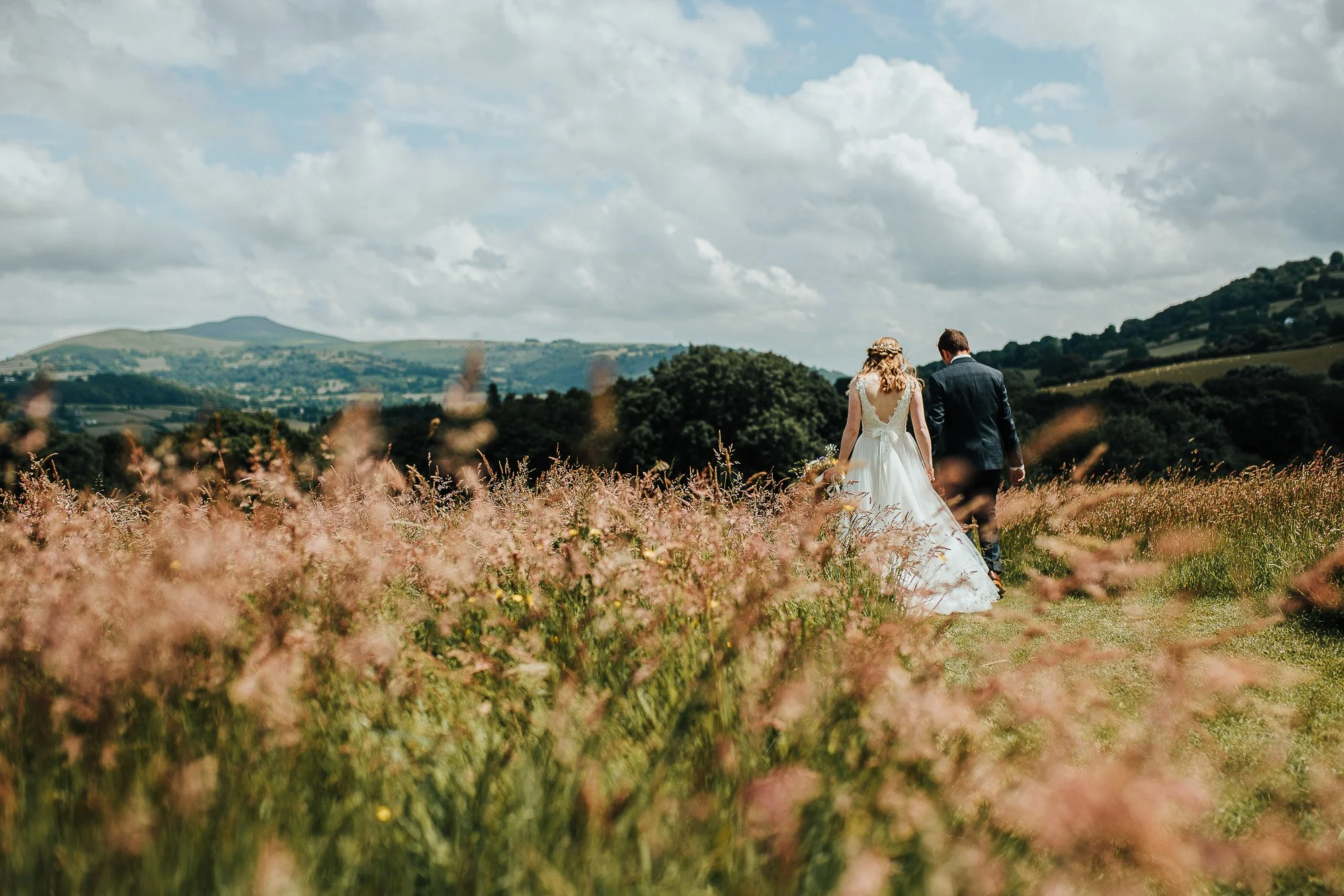 Bride and groom walking through a grassy field with a scenic mountain and cloudy sky backdrop.