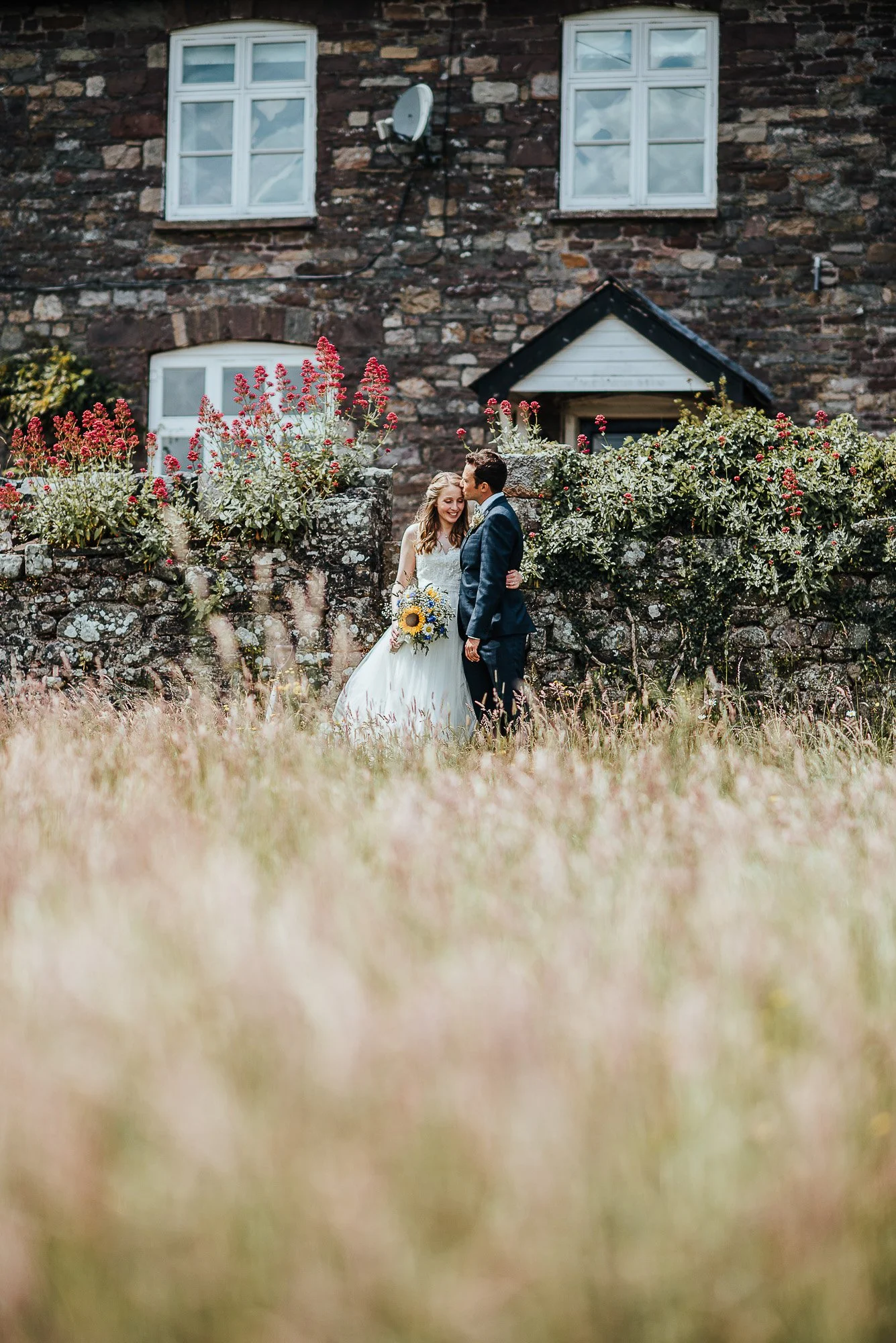 Wedding couple standing in front of stone house with flowers, embracing in a field.