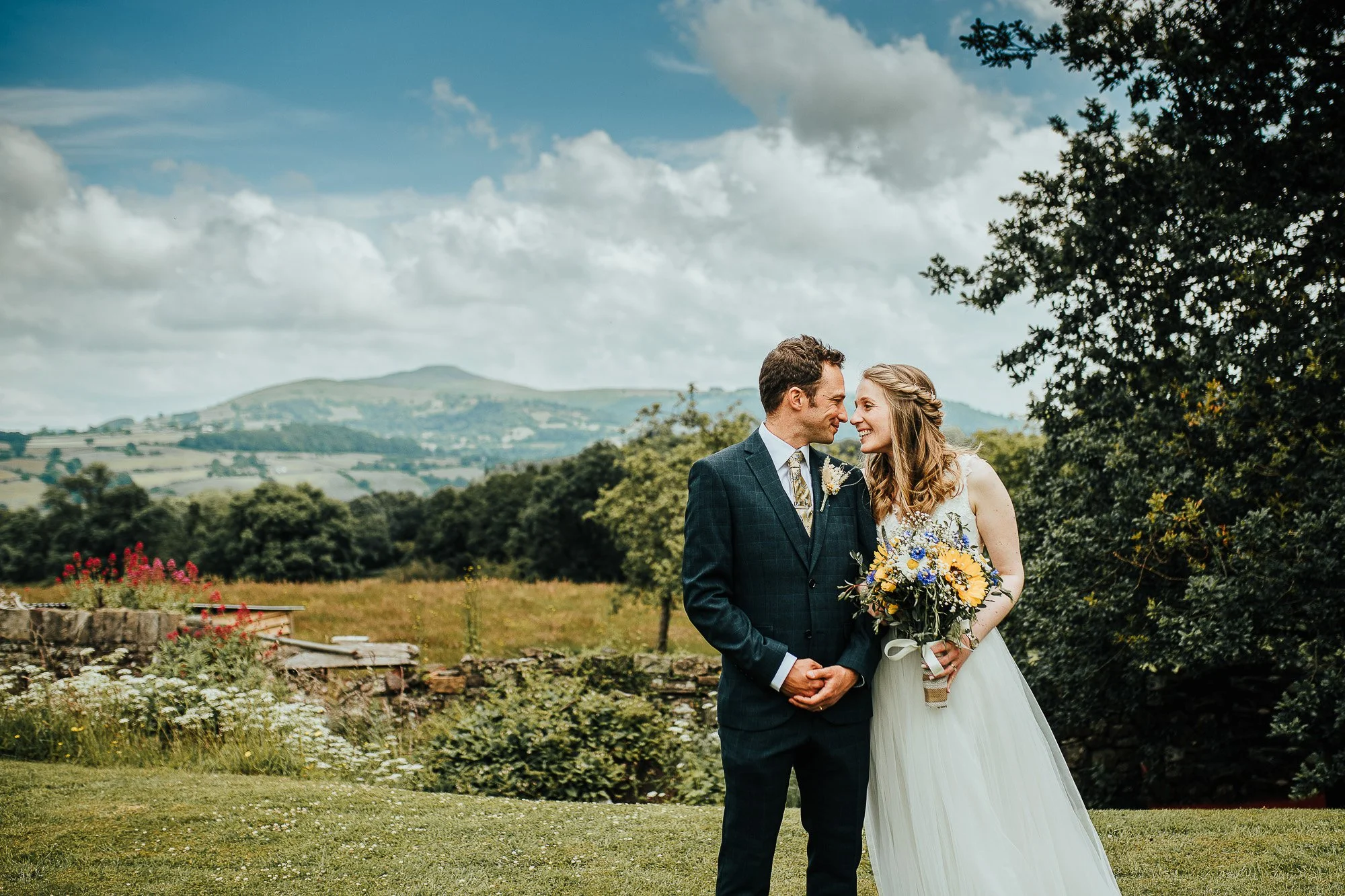 A bride and groom smiling at each other in a scenic outdoor setting with hills and clouds in the background. The bride is holding a bouquet of yellow and blue flowers.