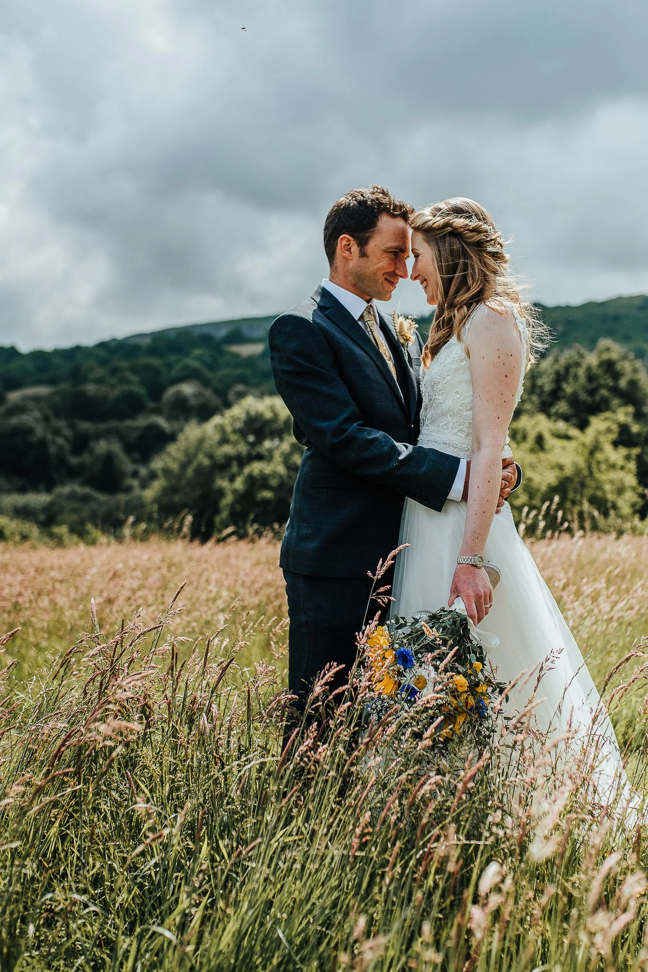 Bride and groom in a field, embracing each other, with the bride holding a bouquet of yellow and blue flowers, against a backdrop of grass and cloudy sky.