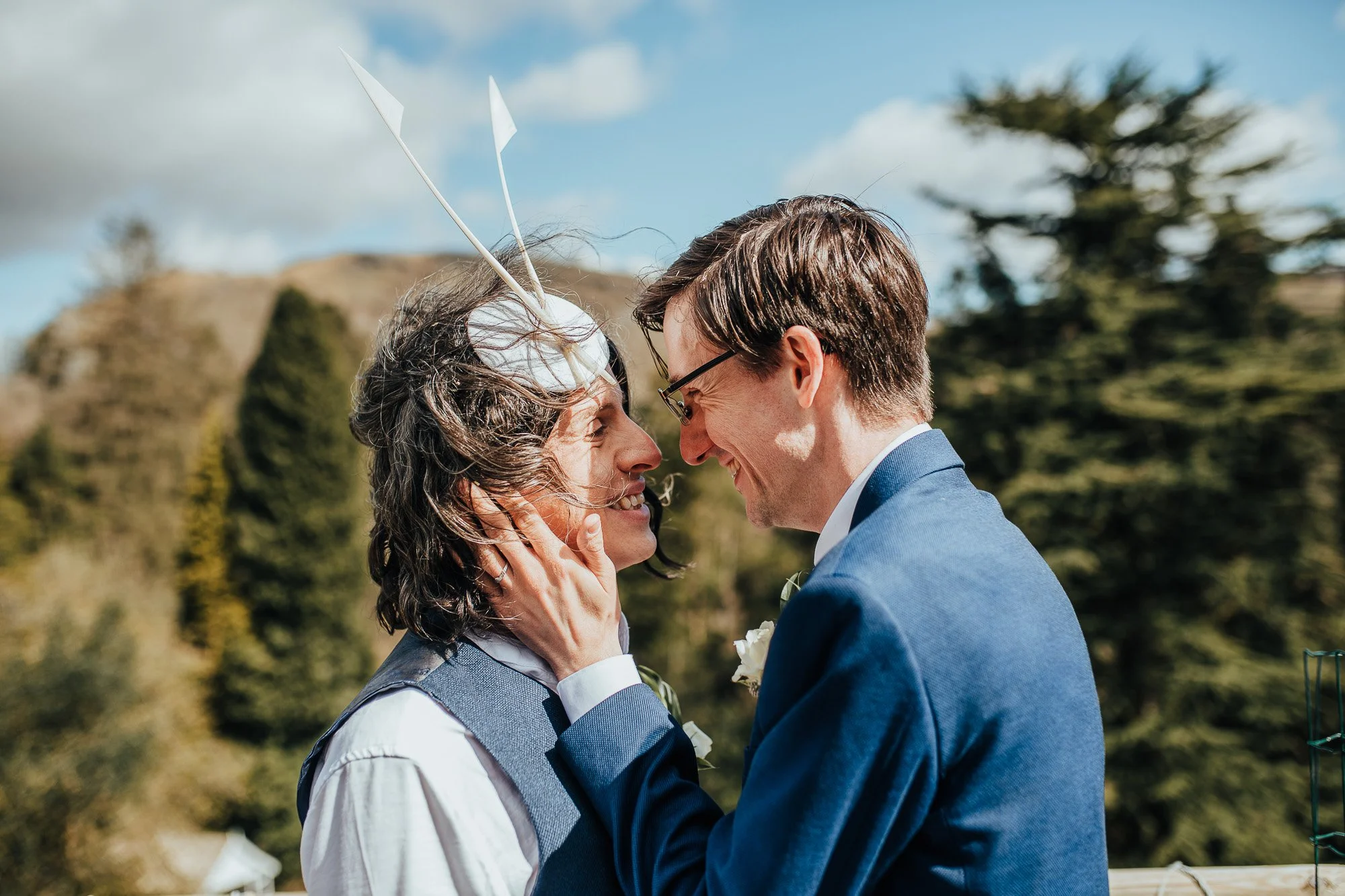Two men in formal attire embracing outdoors, with one wearing a whimsical headpiece, surrounded by nature.