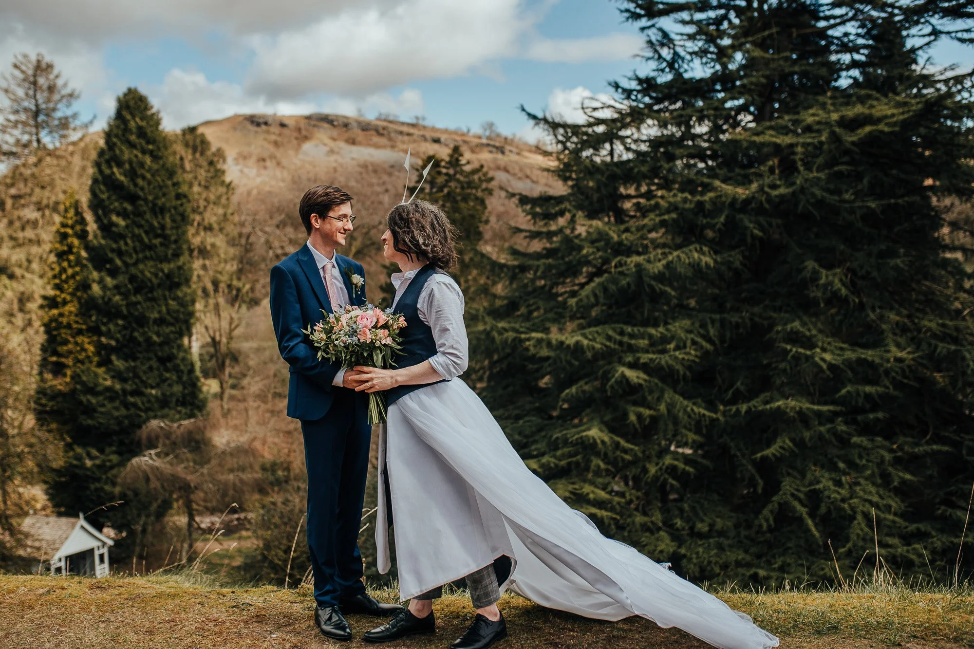 Couple in wedding attire smiling at each other outdoors, surrounded by trees and hills, with one holding a bouquet of flowers.