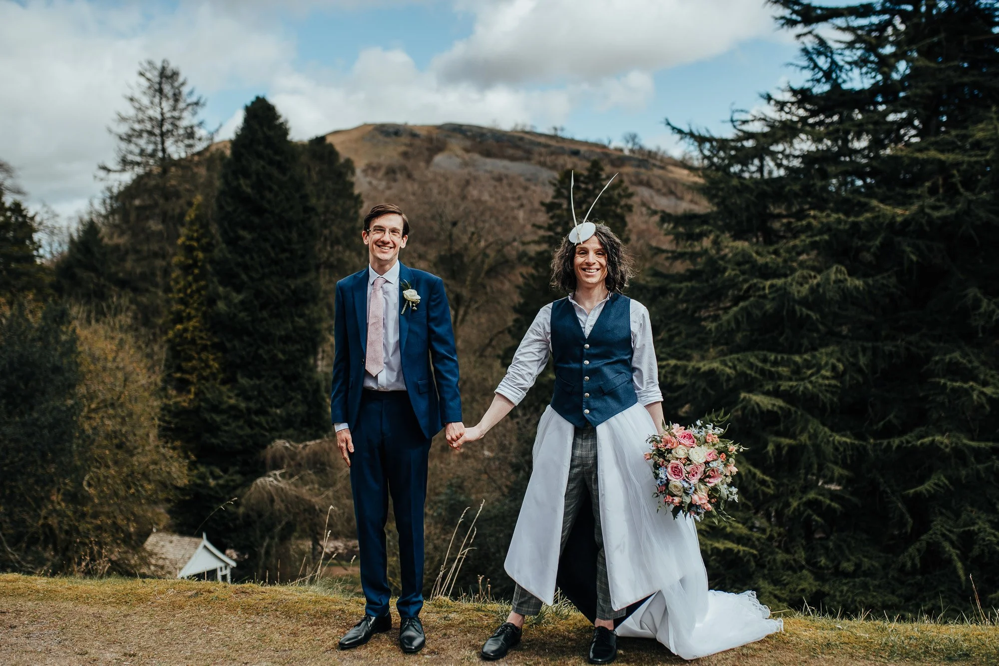 Two people dressed in formal attire, one in a blue suit and tie, the other in a unique outfit with a dress-like train and hat, holding a bouquet of flowers, standing outdoors with trees and hills in the background.