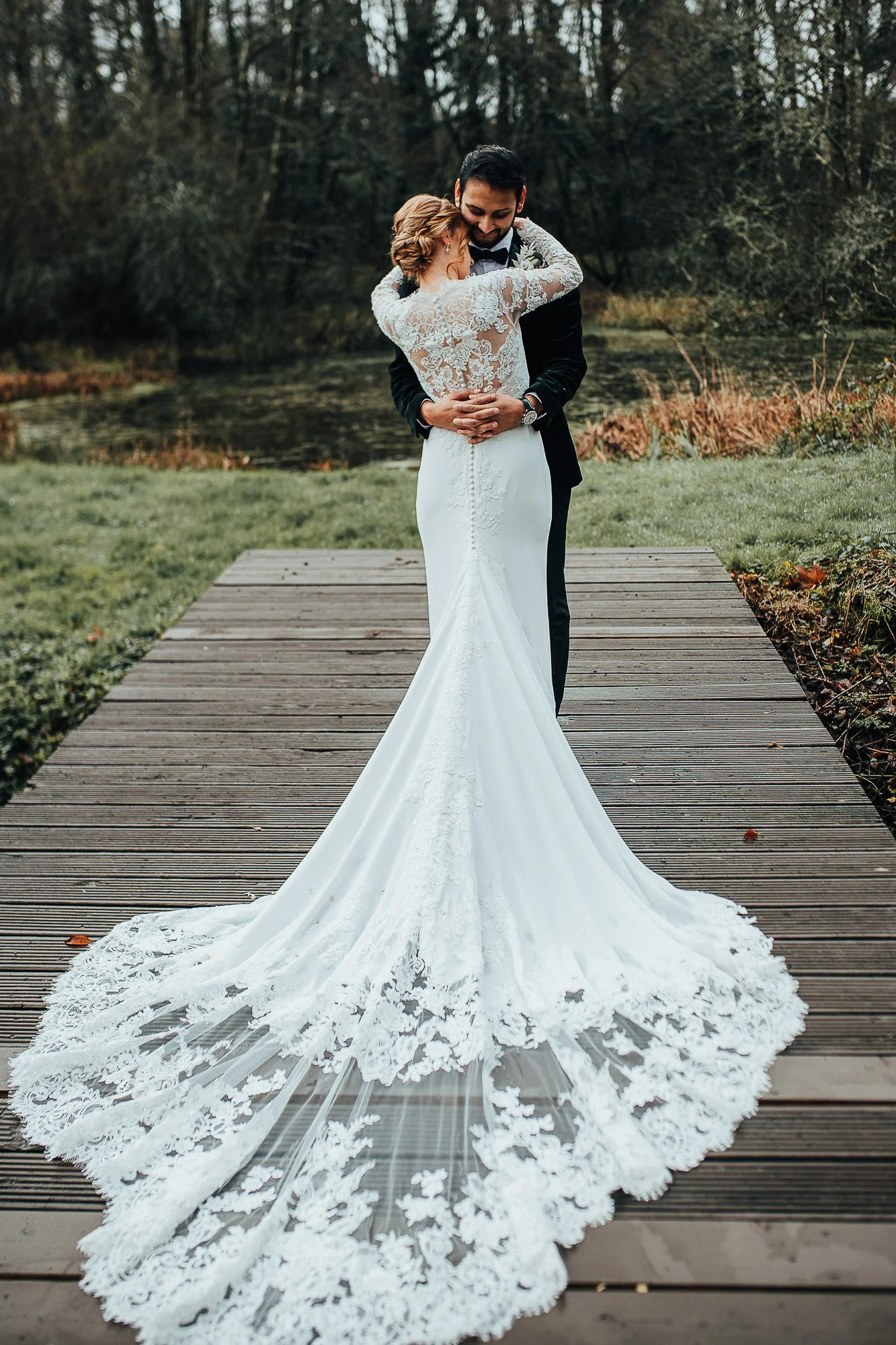 Bride and groom embracing on a wooden path with a scenic natural backdrop. The bride is wearing a long, white lace wedding dress with a detailed train.