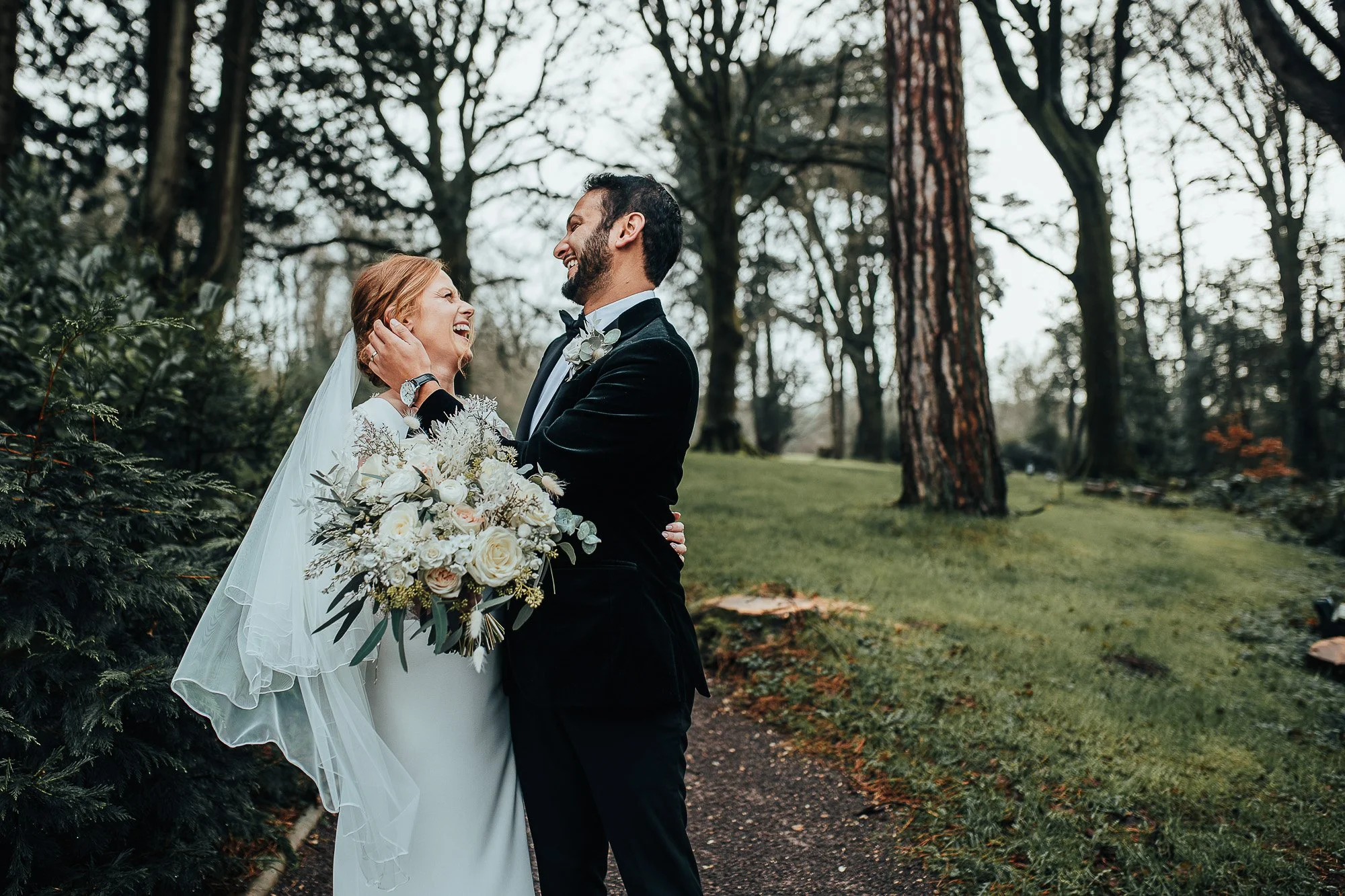 Smiling couple embracing on a wooded path, with the bride holding a floral bouquet.