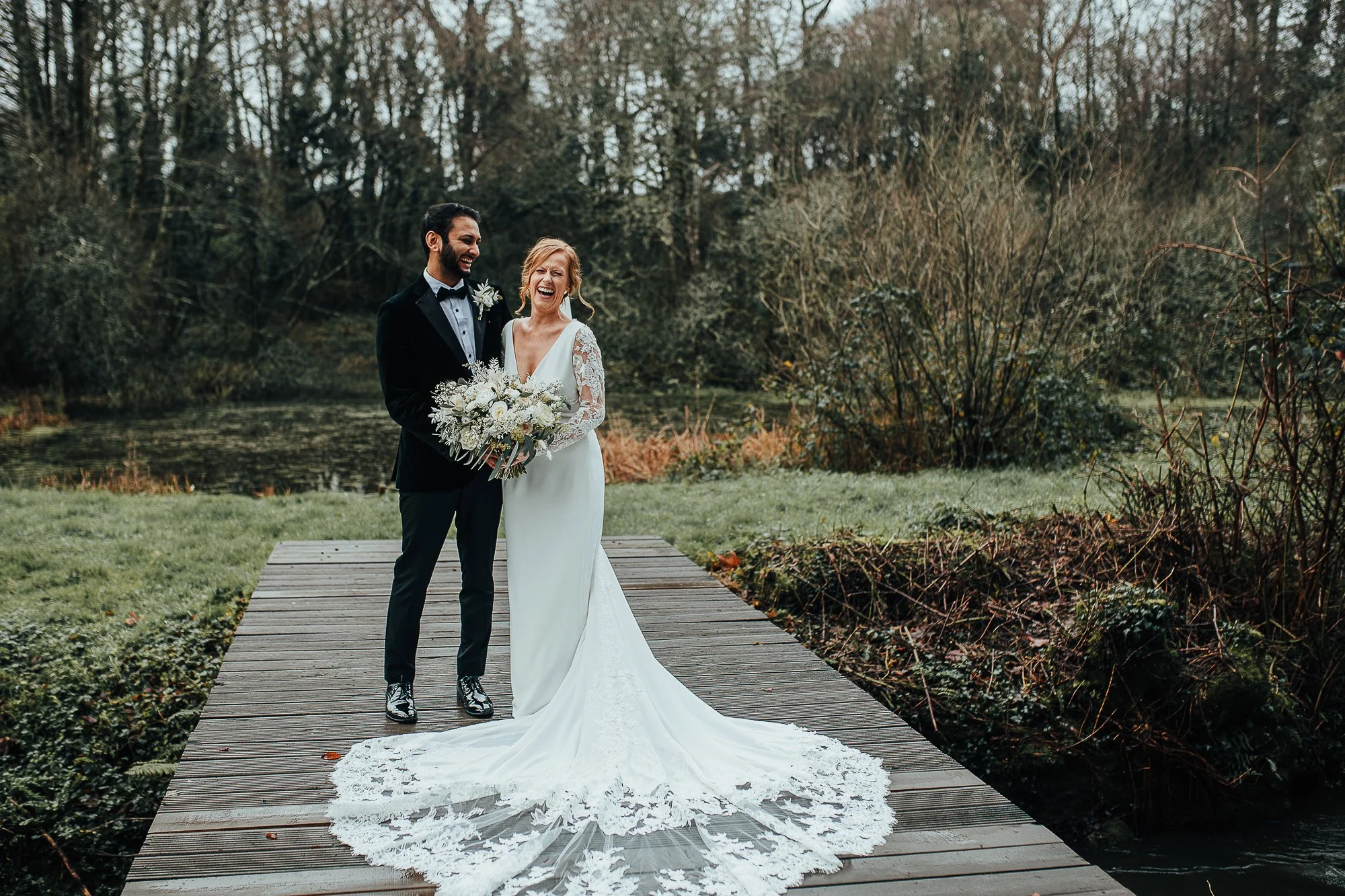 Bride and groom standing on a wooden platform by a pond, surrounded by trees and grass, with the bride in a white dress holding a bouquet.