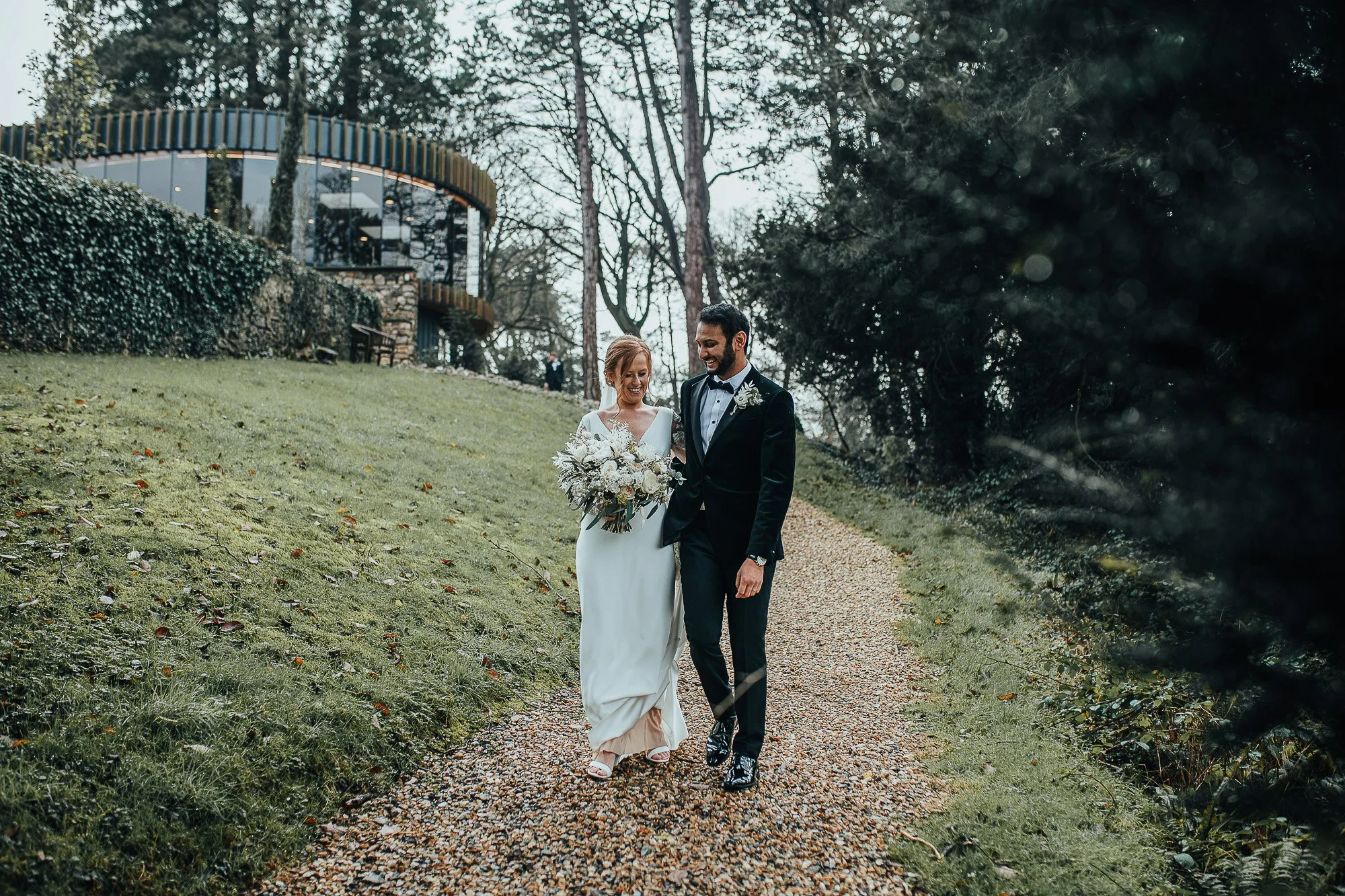 A couple walks hand in hand down a gravel path in a grassy, wooded area. The person on the left is wearing a white wedding dress and holding a bouquet, and the person on the right is in a dark suit. A round building is visible in the background.