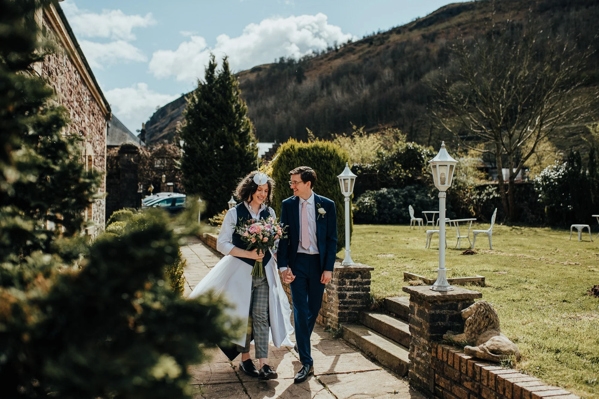 A couple walking hand in hand along a garden path with greenery and outdoor furniture around. The woman holds a bouquet of flowers, and they are both smiling. It is a sunny day with a hilly landscape in the background.