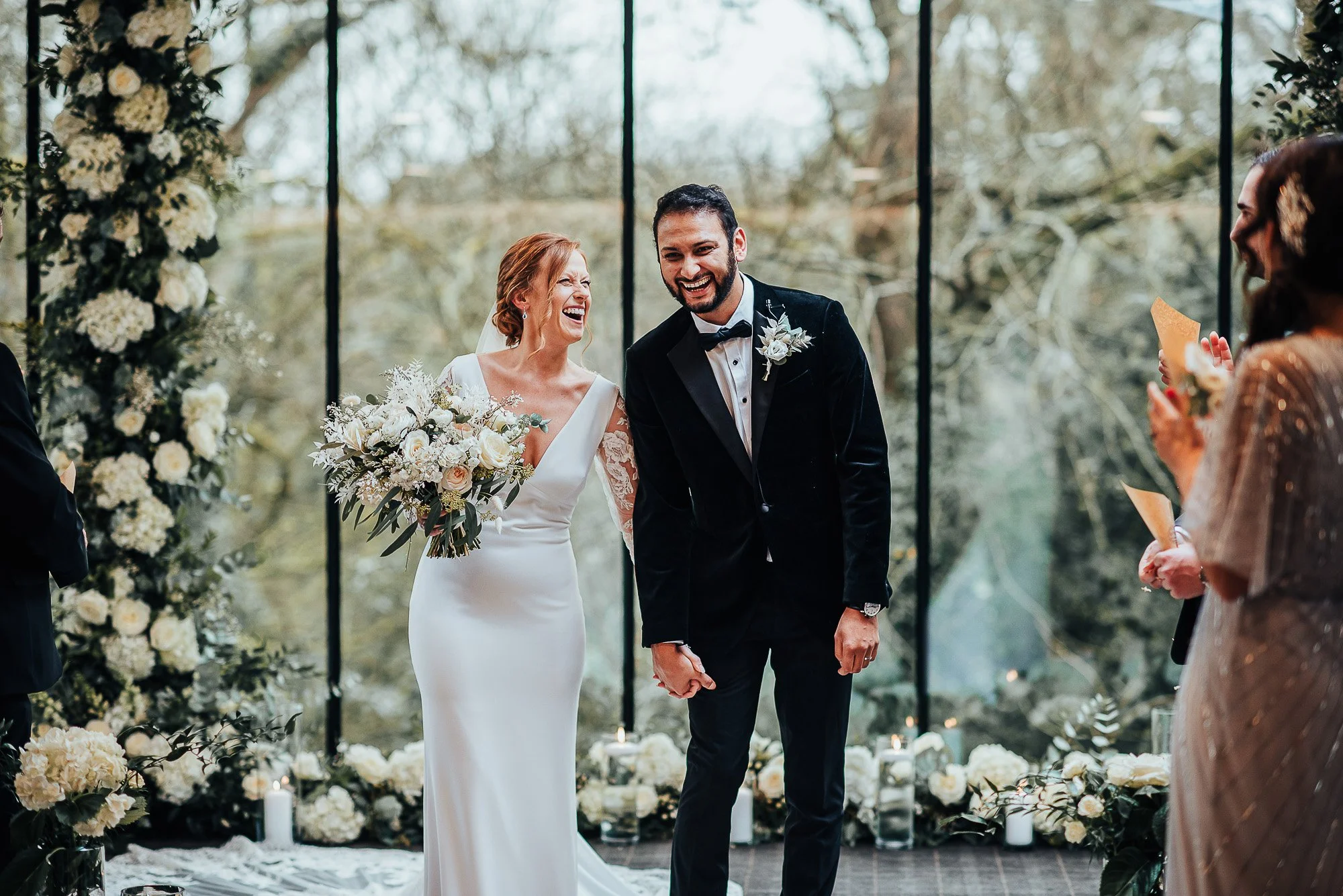 Bride and groom laughing during wedding ceremony with floral arch and guests in background.