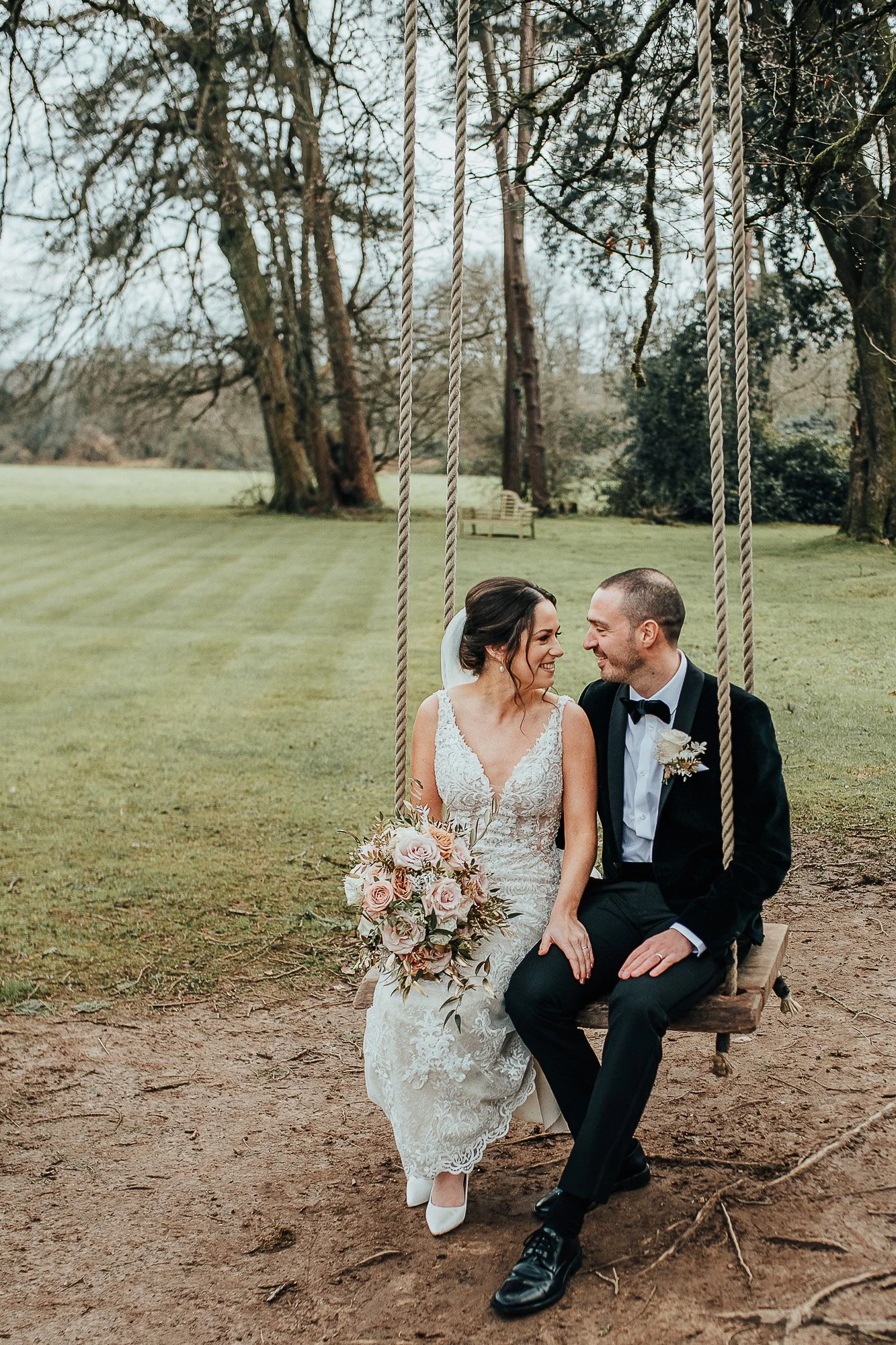 Bride and groom sitting on a swing outdoors, bride holding a bouquet of pink roses, surrounded by trees.