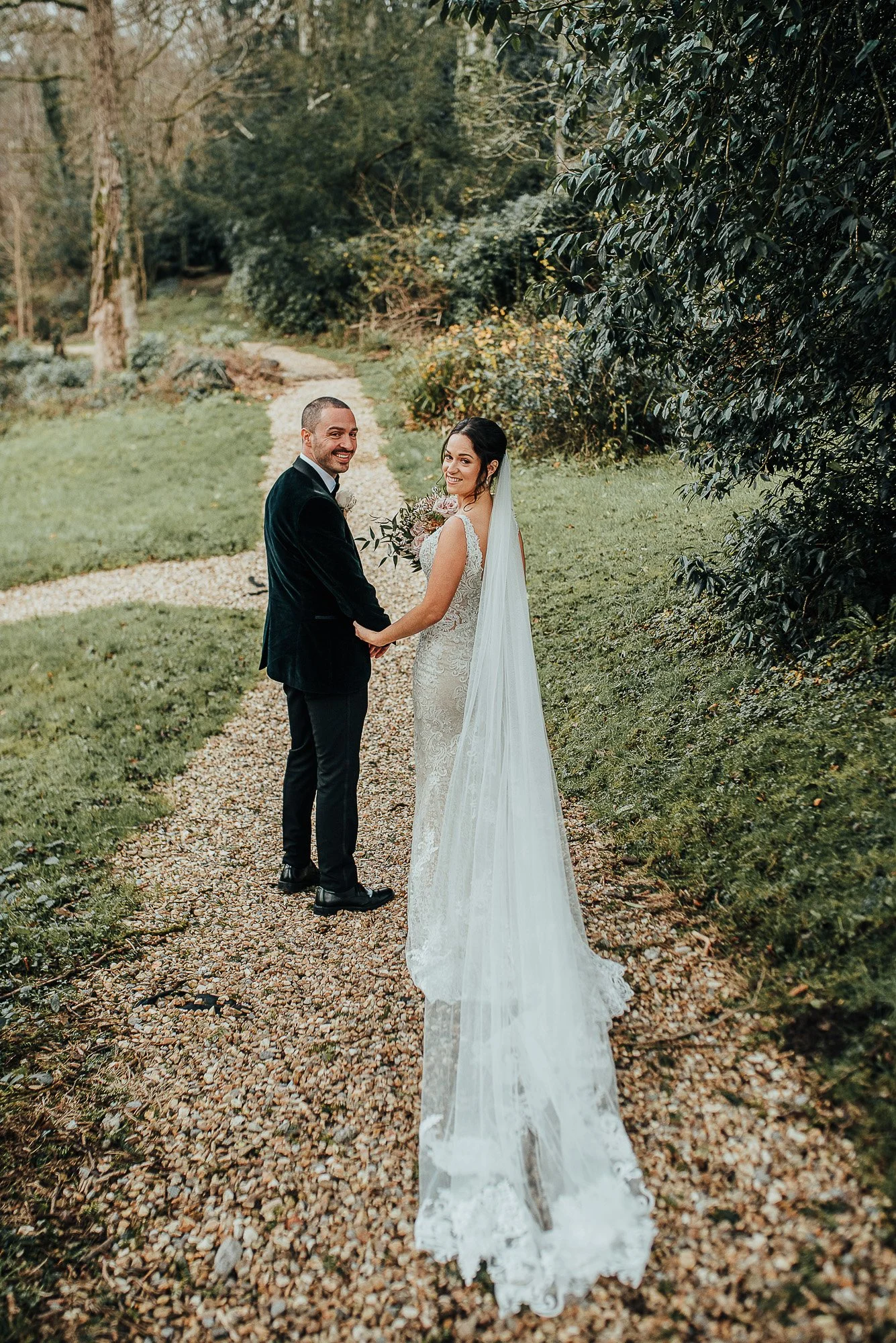 Bride and groom on a pathway in a garden, holding hands and smiling back at the camera. The bride is wearing a lace wedding dress with a long veil, and the groom is in a dark suit. Lush greenery surrounds them.