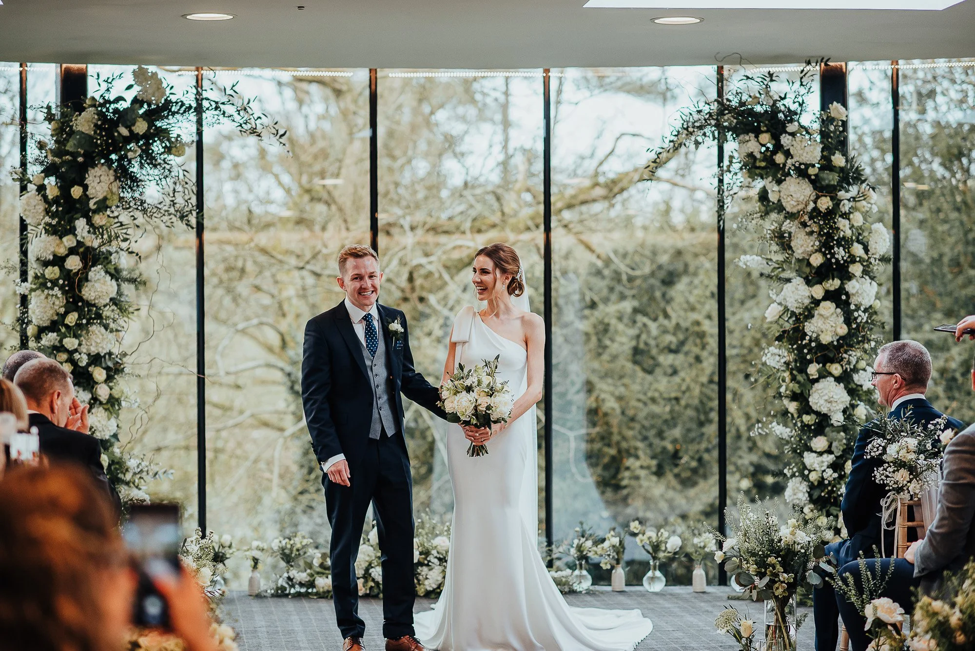 Bride and groom standing at wedding ceremony, holding hands; decorated with flowers.