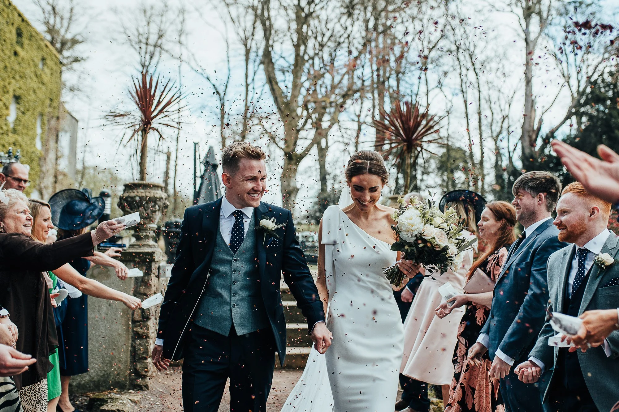 Bride and groom walking hand in hand, surrounded by guests throwing confetti, outdoors at a wedding ceremony.