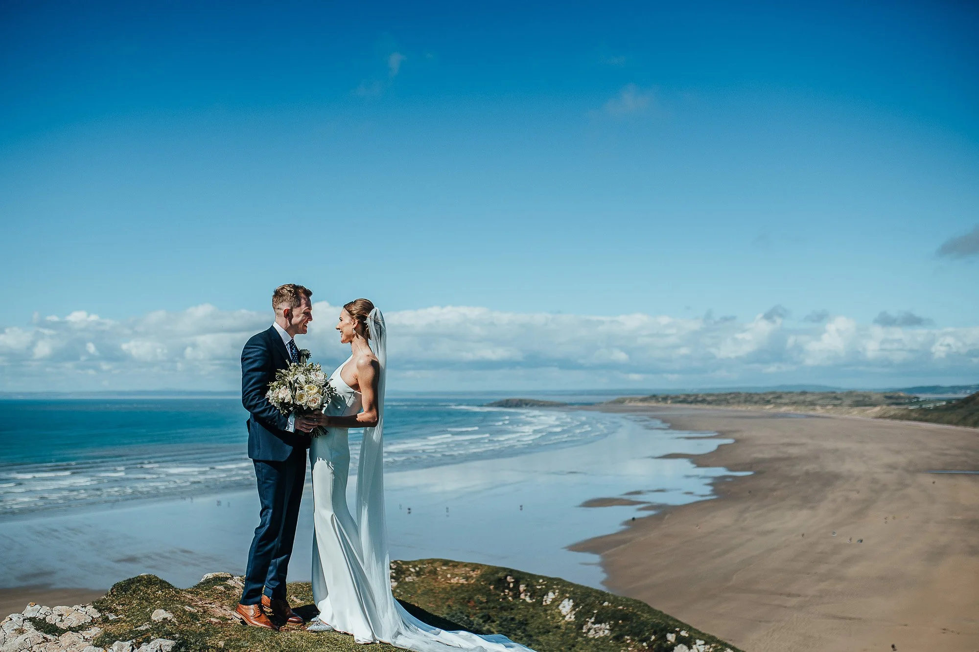 Bride and groom smiling at each other on a cliff overlooking a scenic beach and ocean under a clear blue sky.