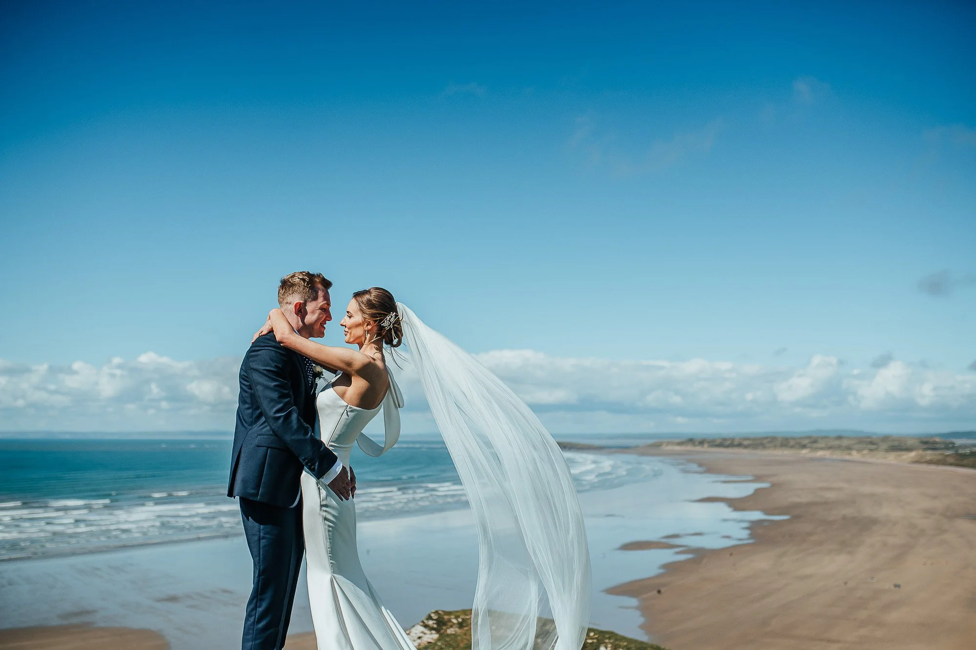Bride and groom embracing on a cliff overlooking the ocean, with a long wedding veil flowing in the breeze, under a clear blue sky.