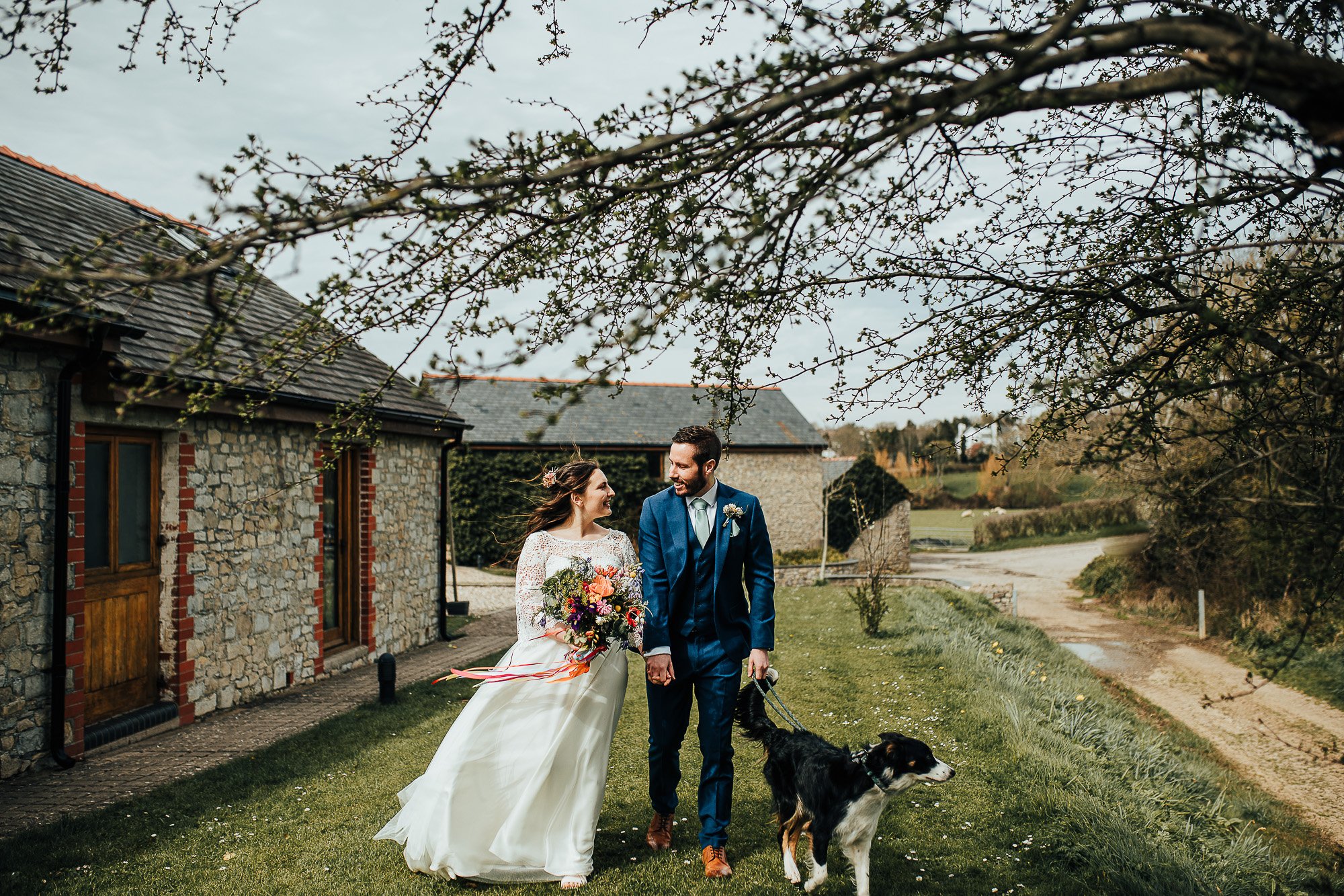 Bride and groom walking with a dog on a grassy path near stone buildings, under a tree with budding branches.