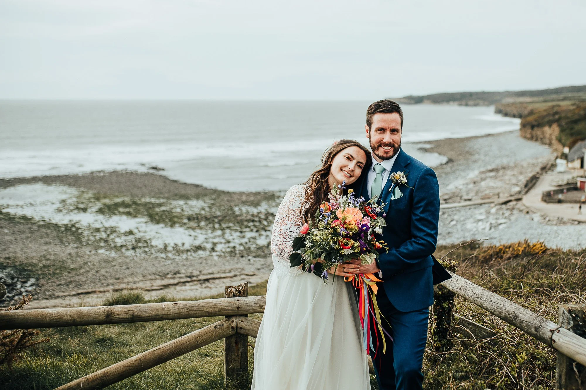 Bride and groom standing outdoors near a coastal landscape, smiling and holding a colorful bouquet, with the ocean in the background.