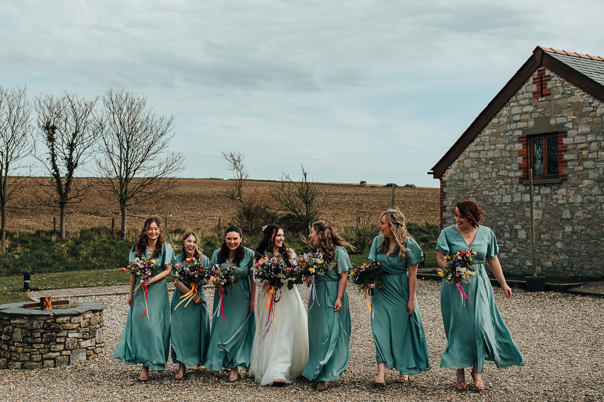 Group of bridesmaids in teal dresses holding bouquets walking outdoors near a stone building.