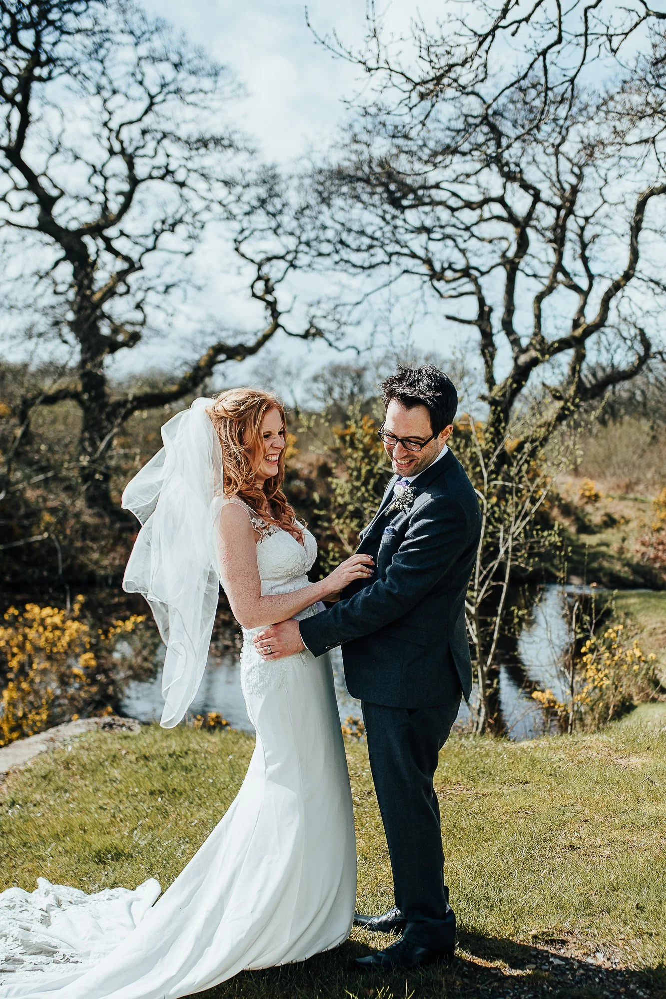 Happy couple standing outdoors in wedding attire, bride in a white dress and veil, groom in a dark suit, with a scenic background of trees and a small stream.