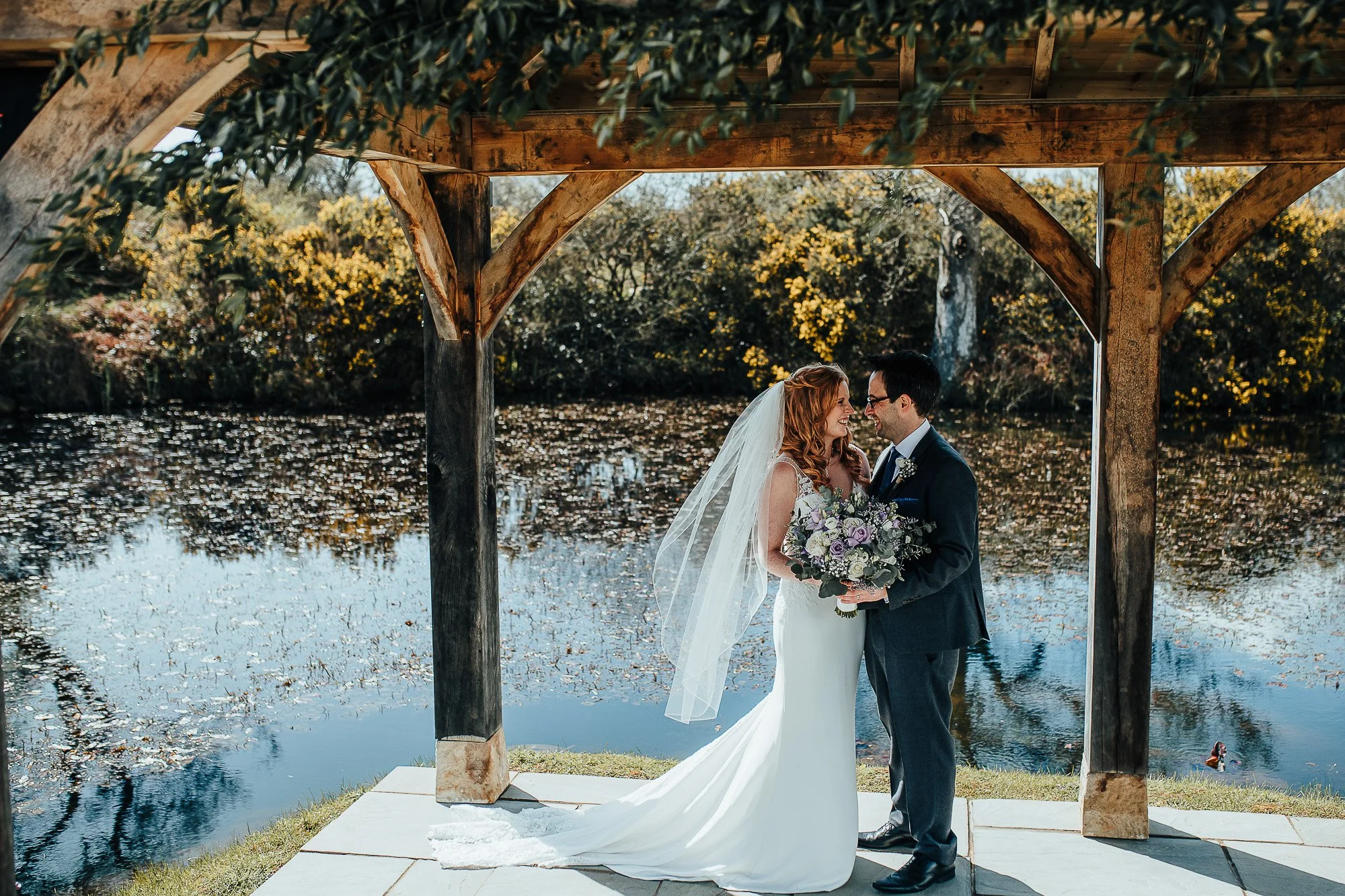 Bride and groom embracing under wooden arch near pond, with bride holding bouquet and veil flowing, surrounded by greenery.