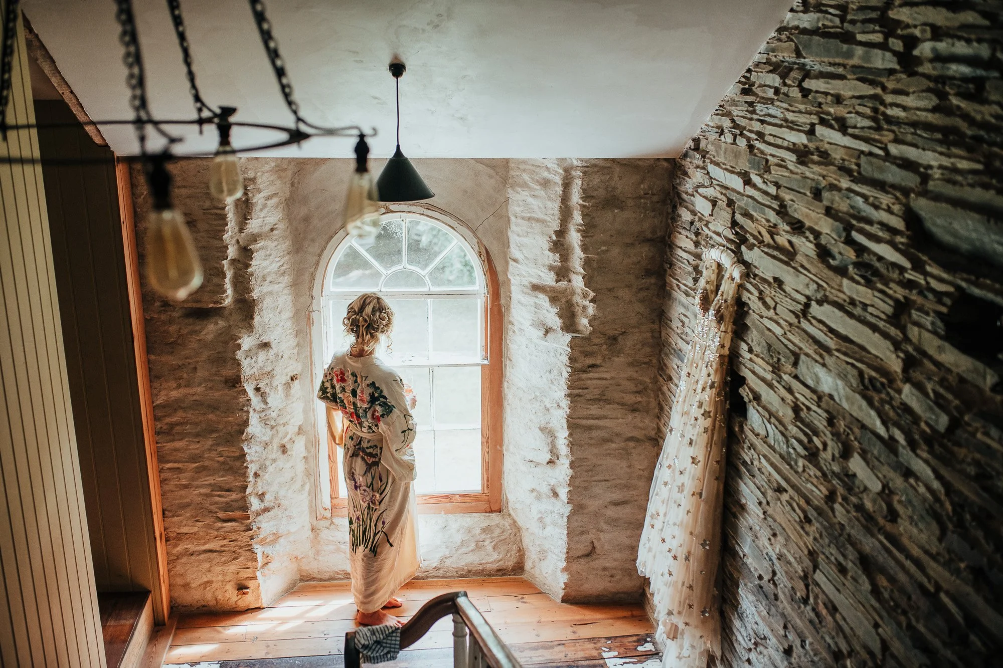 Woman in floral robe standing by arched window in rustic stone-walled room with hanging light fixtures.