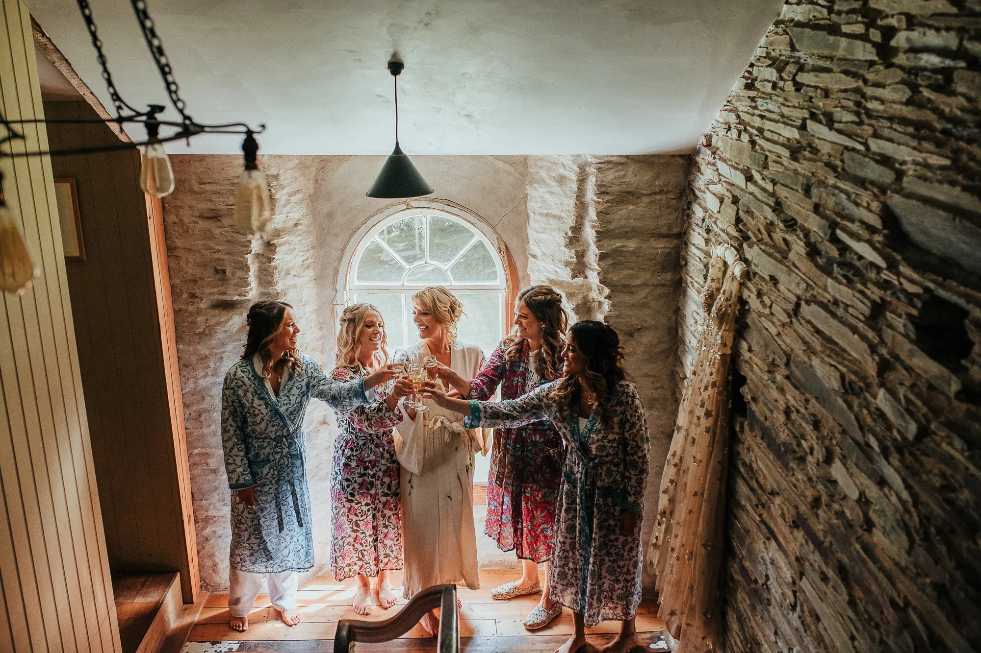 A group of women in colorful robes toast with champagne glasses in a stone-walled room, under a hanging light fixture, next to a window with a view.