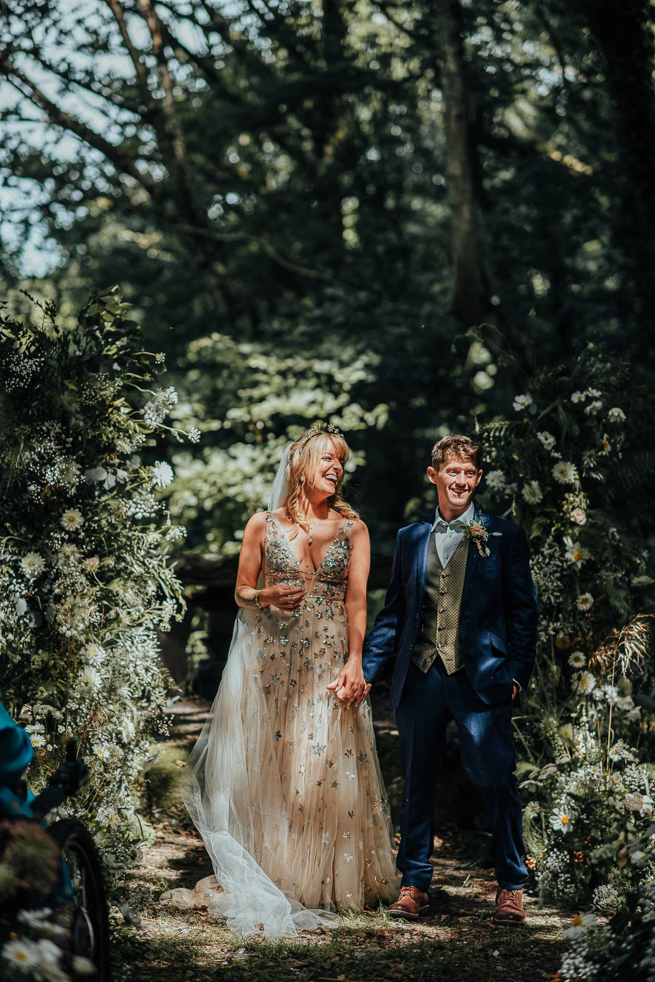 Bride and groom standing outdoors in a forest, surrounded by greenery and flowers, wearing a floral wedding dress and a suit.