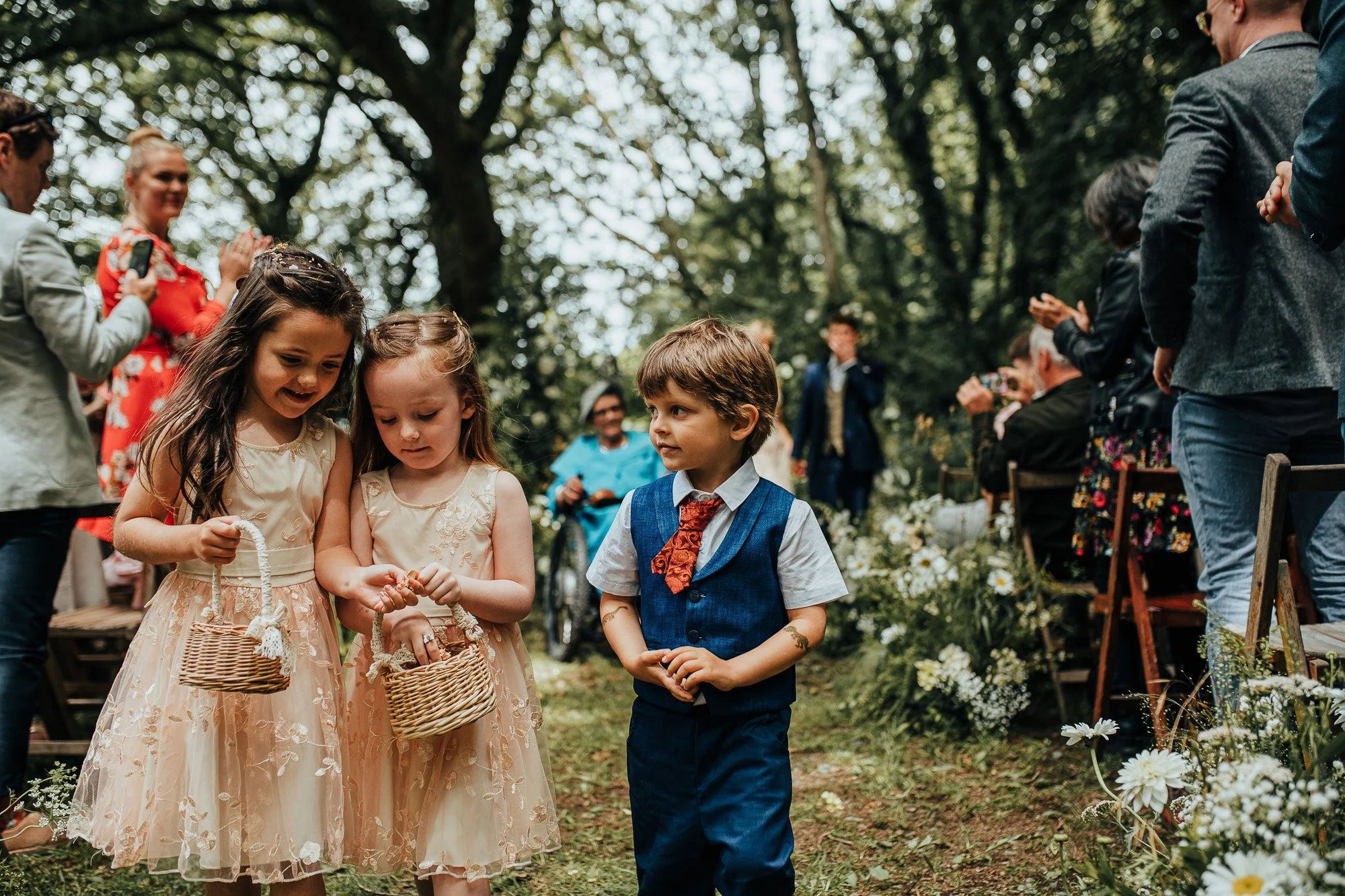 Two young girls in matching lace dresses hold woven baskets and walk with a boy in a vest and tie at an outdoor wedding ceremony. Guests are seated and clapping in the background. Flowers line the aisle in a wooded area.
