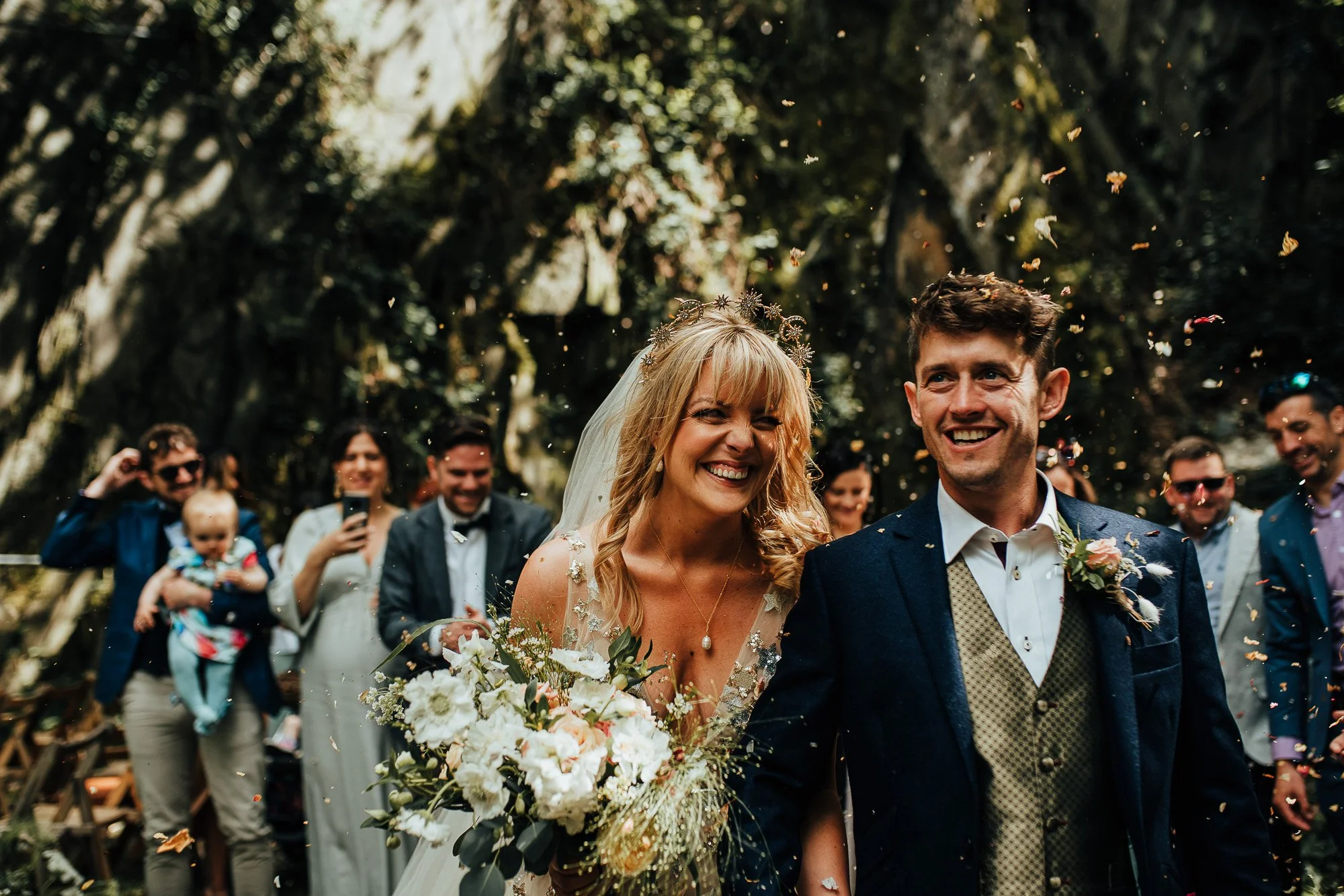 Smiling bride and groom walking down an outdoor aisle with guests throwing confetti, surrounded by trees and greenery.