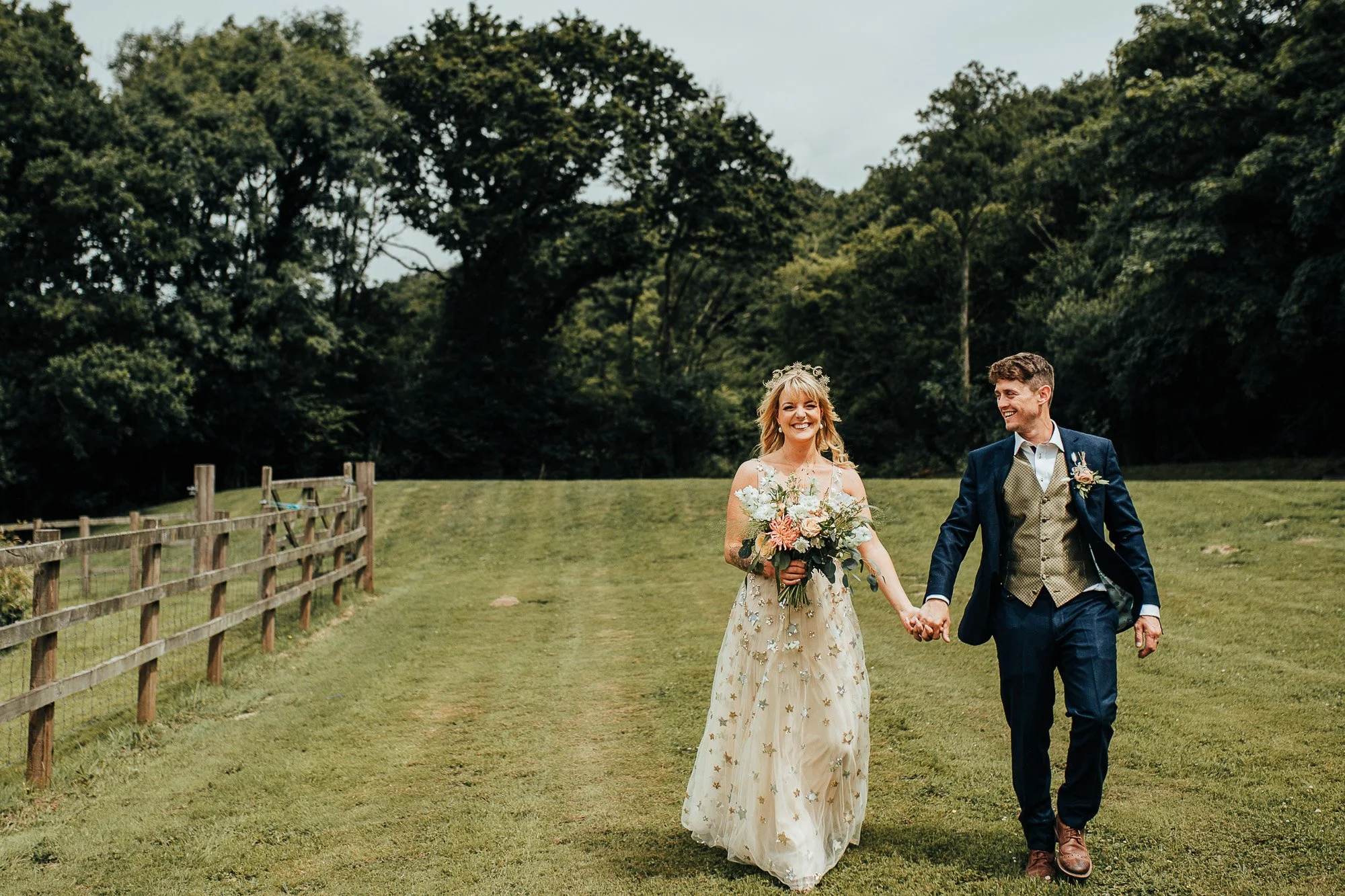 Bride and groom walking hand-in-hand in a grassy field with a wooden fence and trees in the background, bride holding a bouquet.