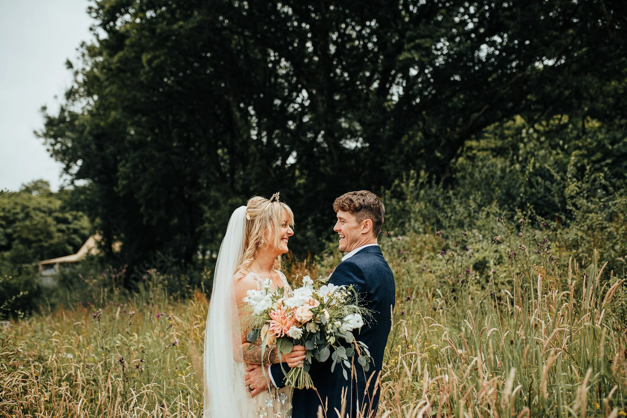Bride and groom smiling at each other holding a wedding bouquet in a field with tall grass and trees in the background.