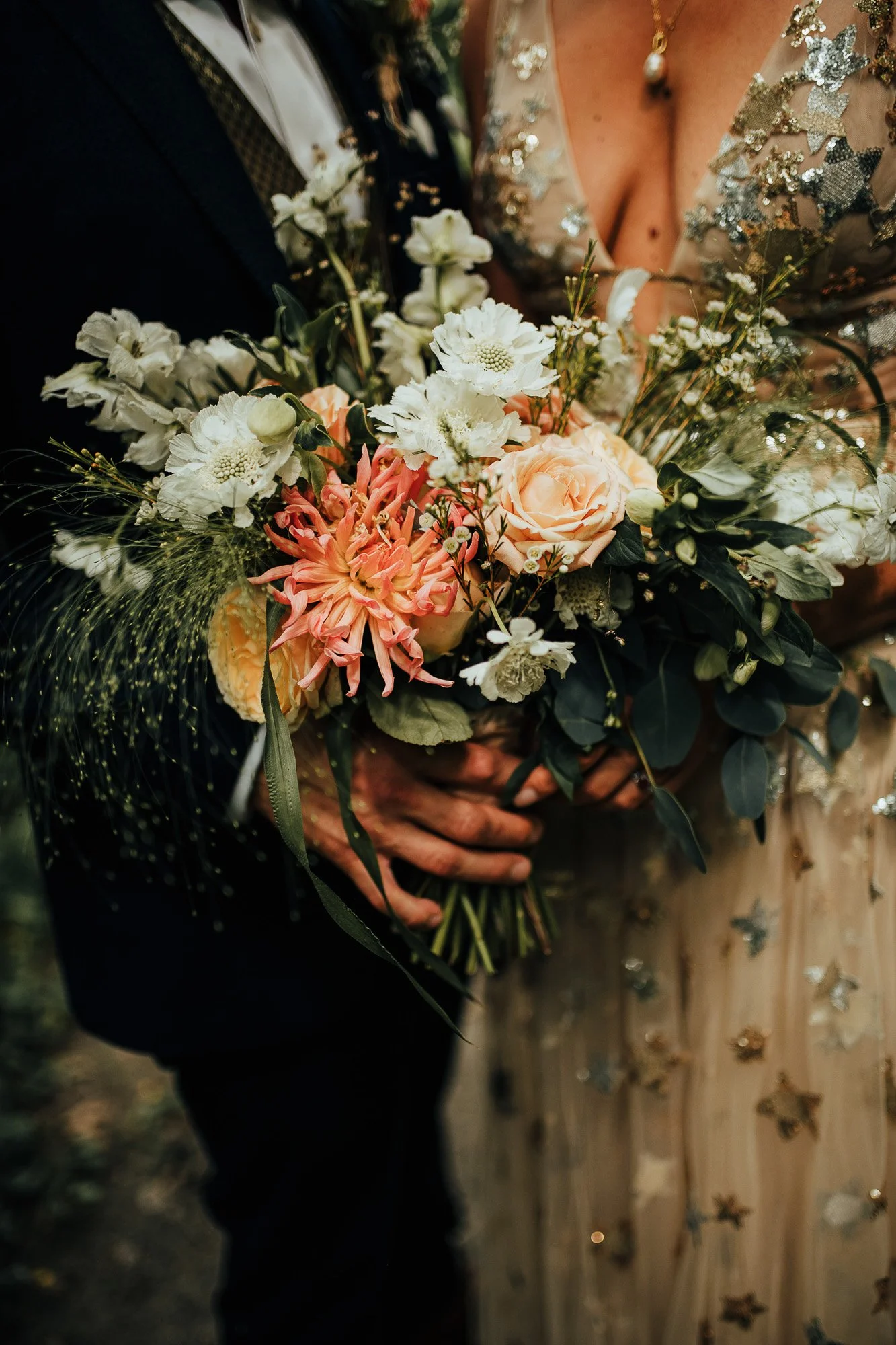 Bride holding bouquet with peach and white flowers, greenery; close-up, wedding dress detail.