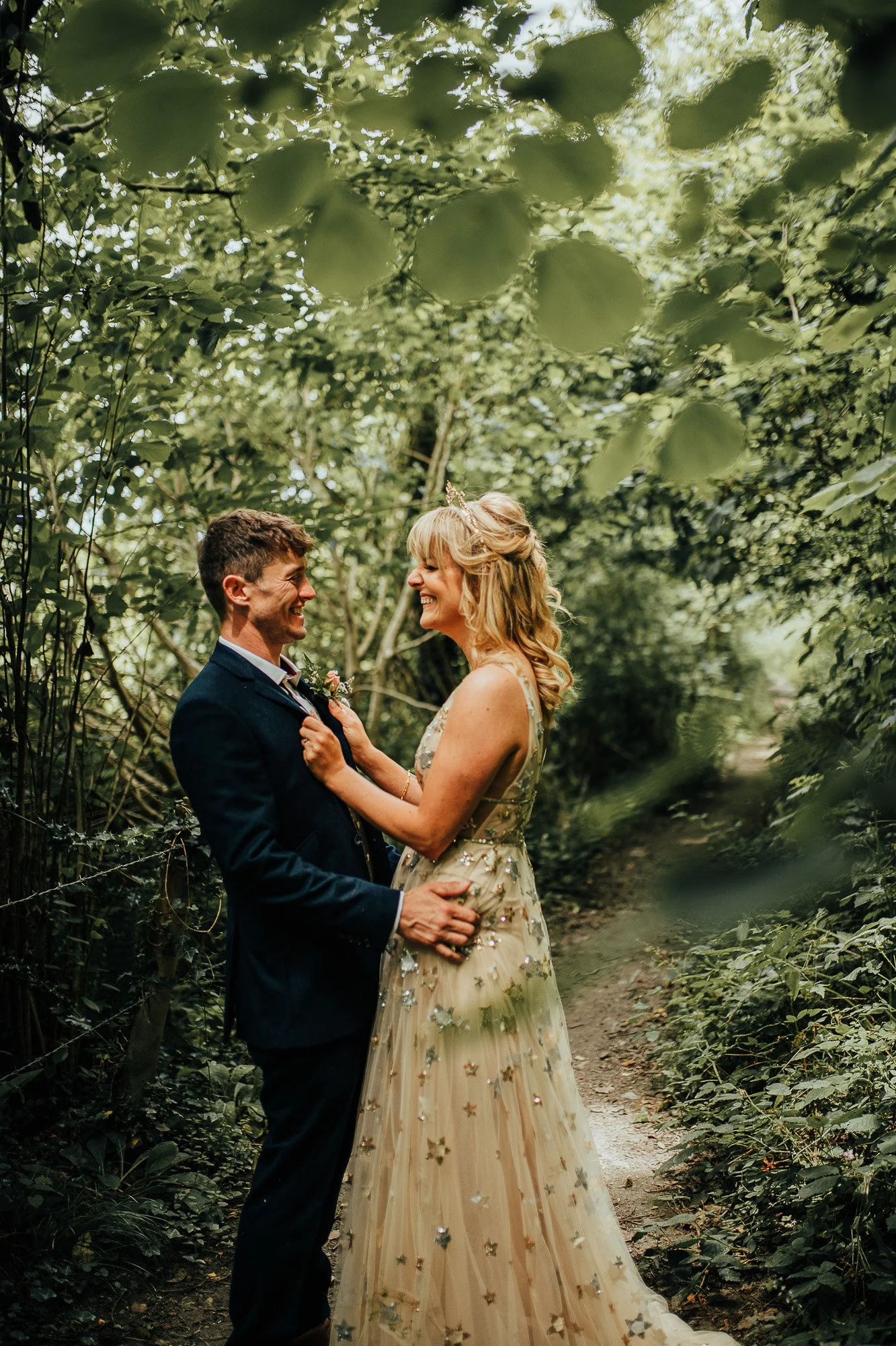 A couple standing together, smiling at each other in a wooded path, surrounded by greenery. The woman is wearing a decorative gown, and the man is in a suit.
