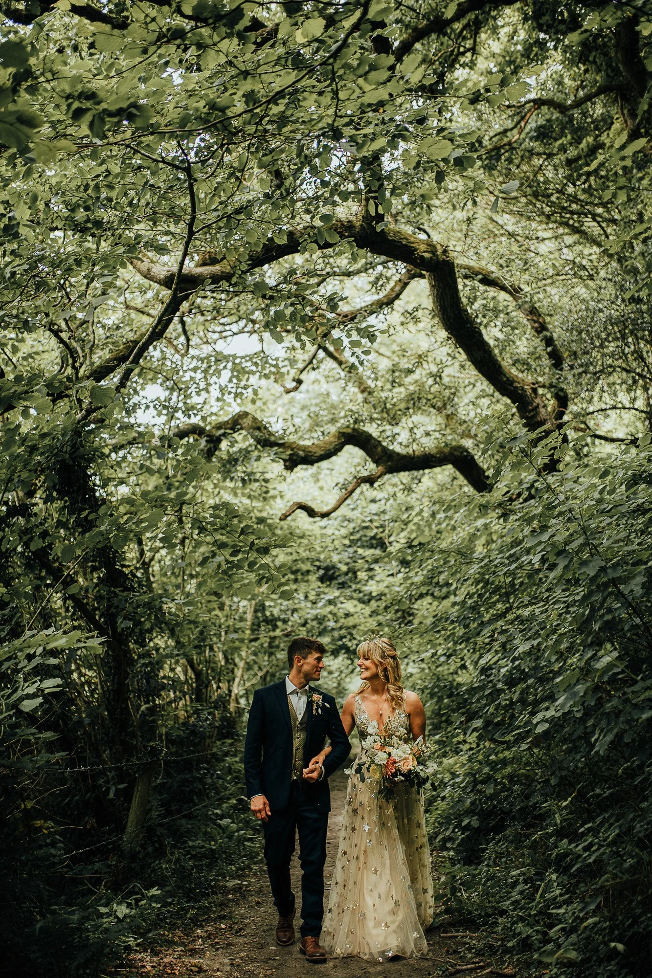 A couple in wedding attire walking through a lush green forest path, with dense foliage overhead. The bride is holding a bouquet and wearing a gown, while the groom is in a suit.