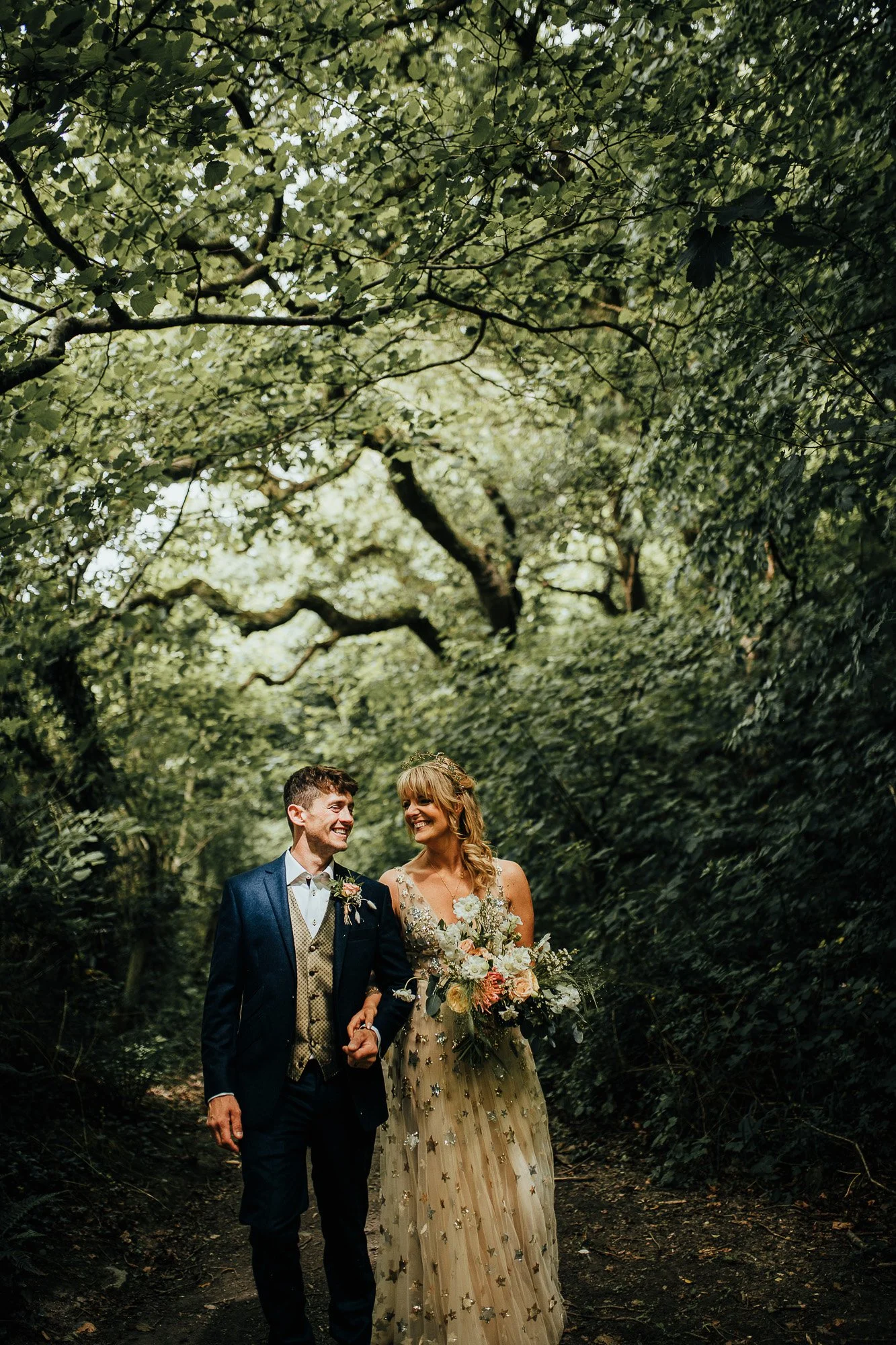 Bride and groom walking through a lush green forest, smiling and holding hands; bride wearing a floral dress and groom in a suit.