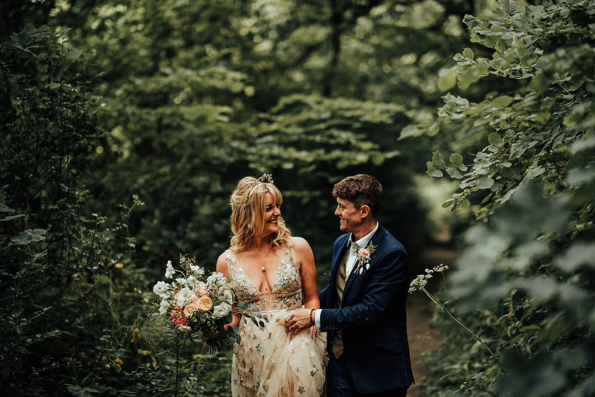 Wedding couple in a forest path, bride holding a bouquet and wearing a floral dress, groom in a navy suit.