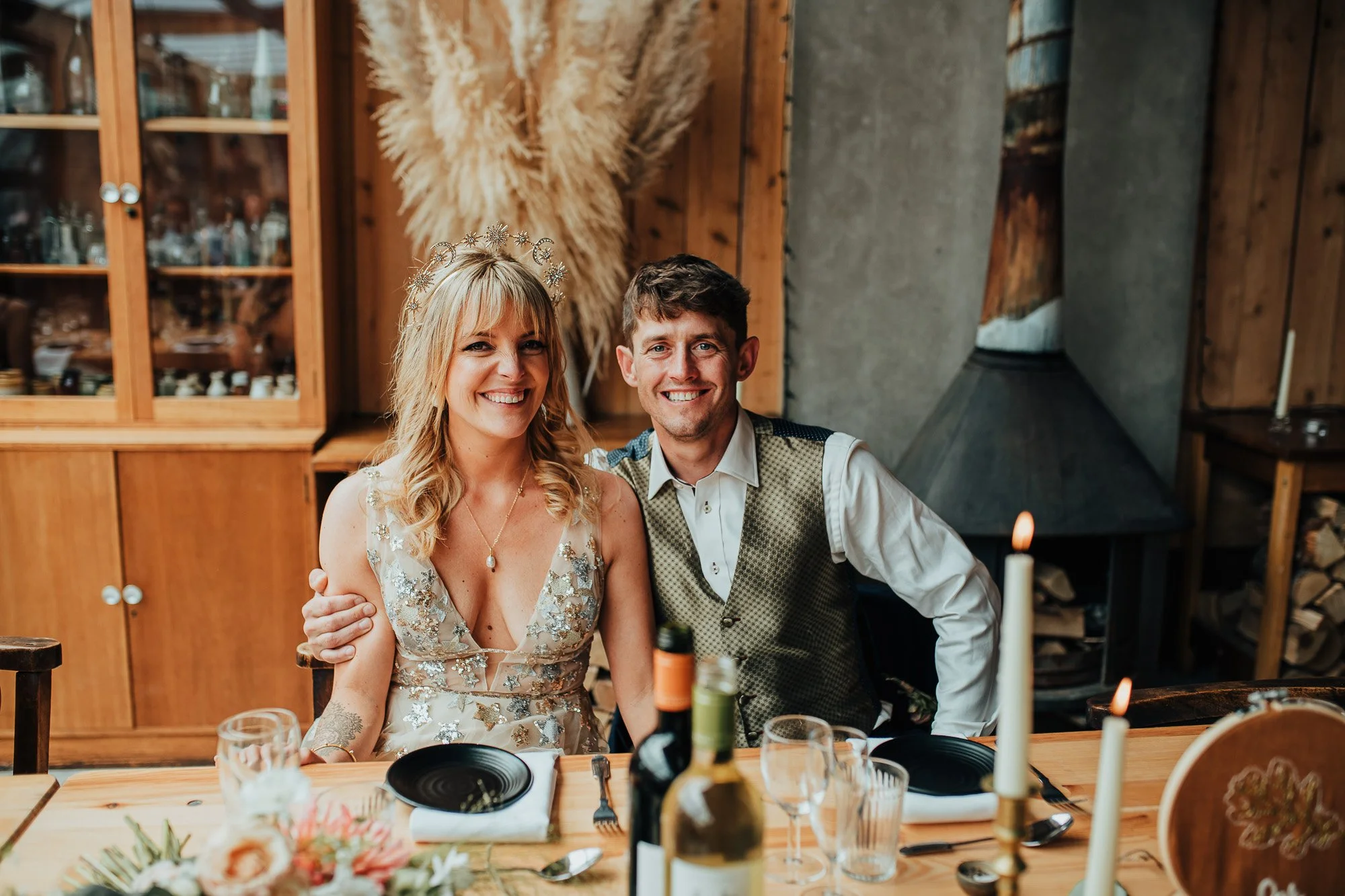 A couple smiling and seated at a table, dressed in formal attire. The table has wine bottles, glasses, and decorative items. The background includes wooden cabinets and dried pampas grass.