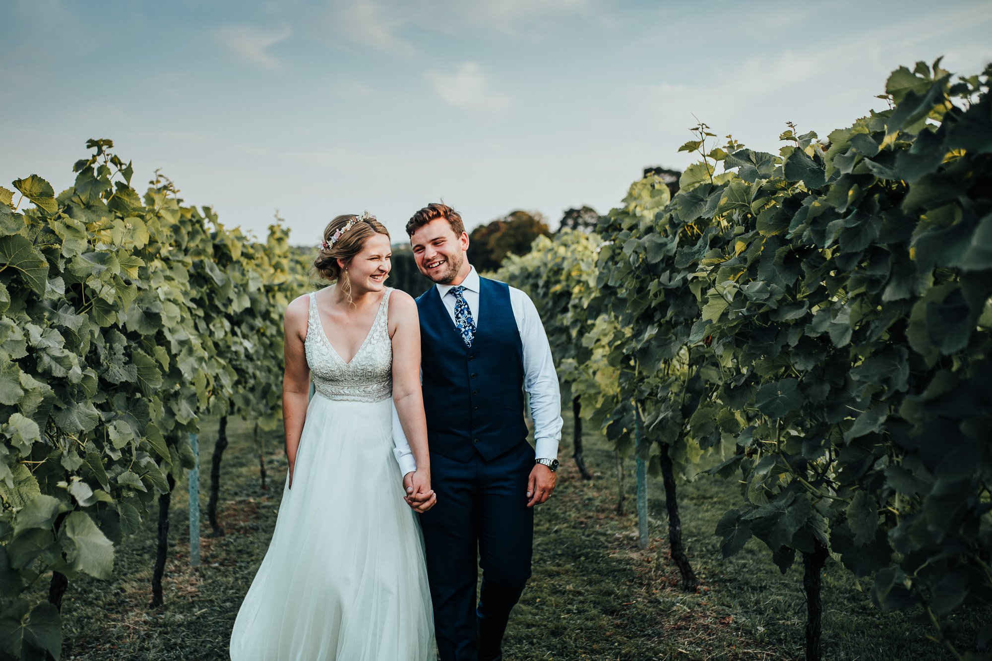 Bride and groom walking together in a vineyard, smiling and holding hands. The bride wears a white dress and floral headpiece, the groom wears a navy suit and tie.