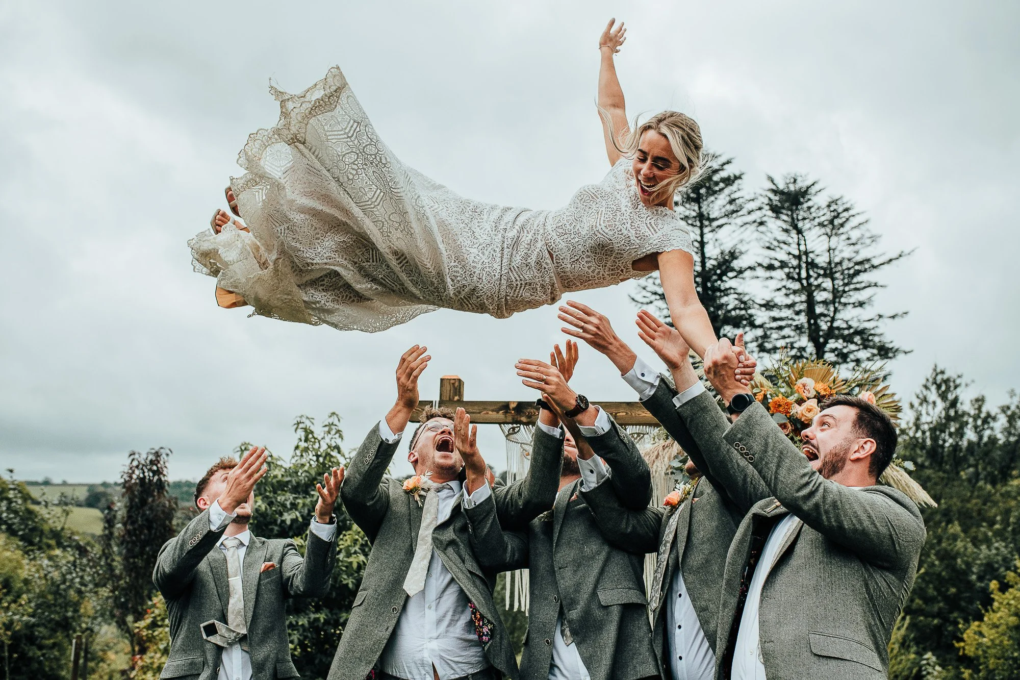 Bride being joyfully tossed in the air by groomsmen outdoors, everyone smiling, with trees and cloudy sky in the background.