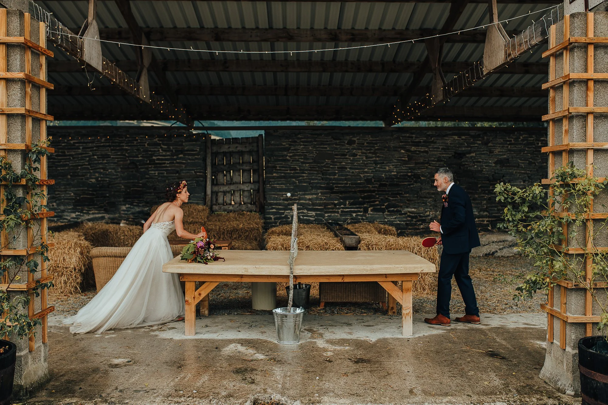 Bride and groom playing ping pong in a rustic barn setting, decorated with string lights and hay bales, during their wedding reception.