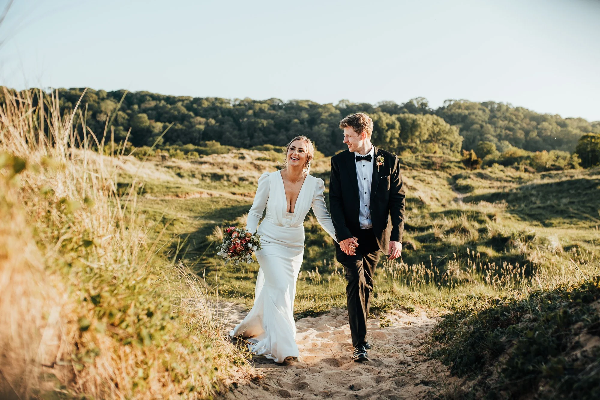 Bride and groom walking on a sandy path in a grassy landscape