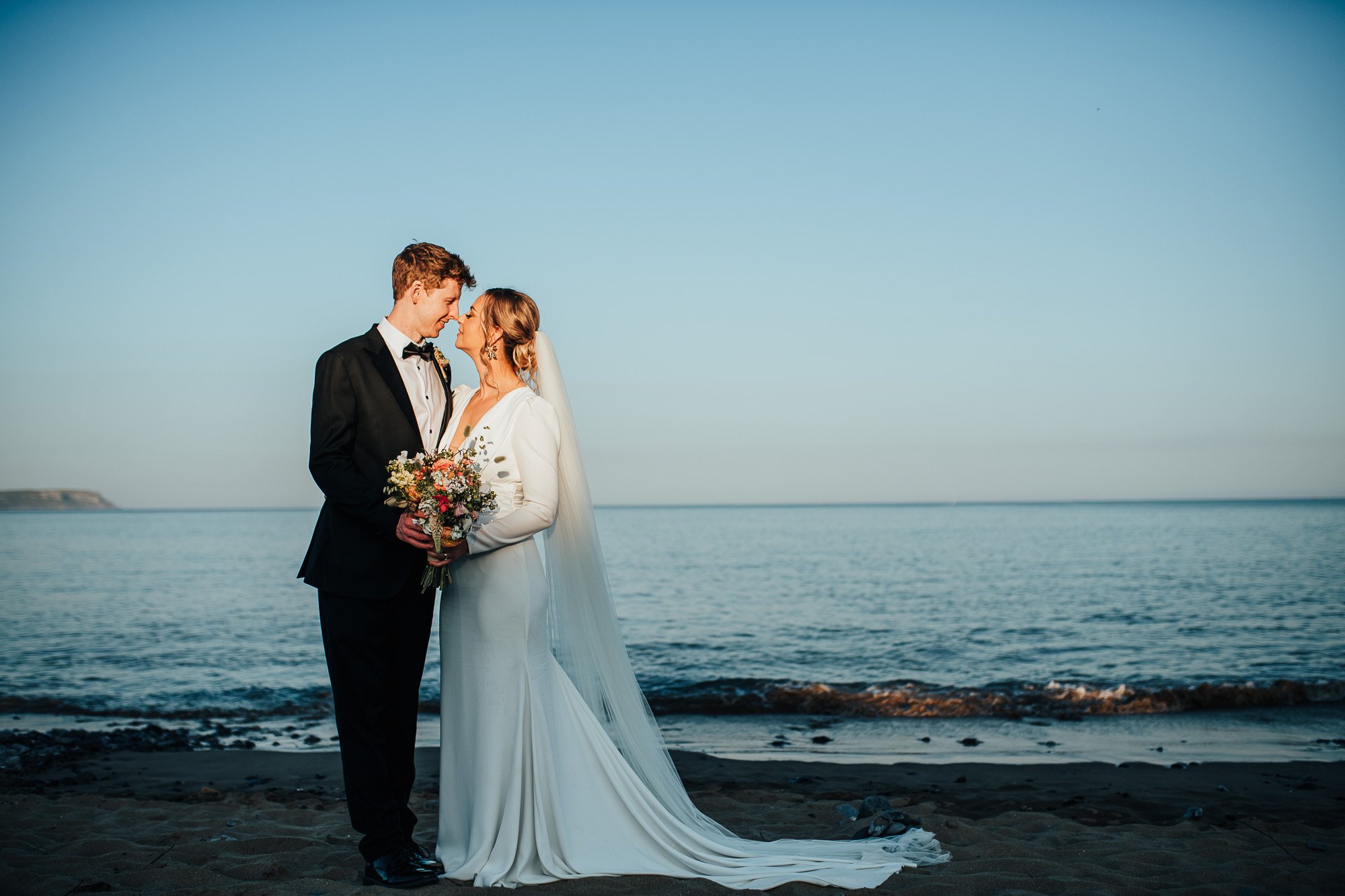 Bride and groom standing on a beach, groom in a black tuxedo, bride in a white gown holding a bouquet, ocean in the background, clear sky.