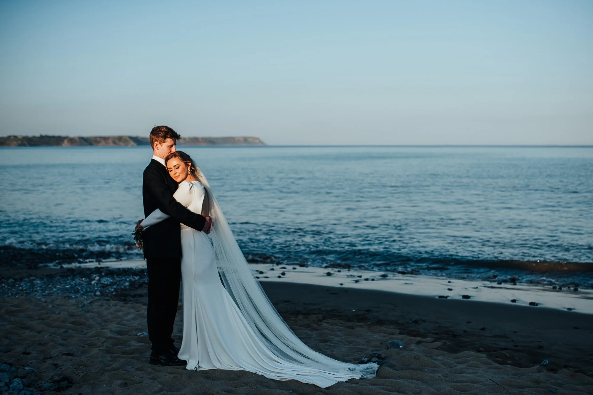 Bride and groom embracing on a beach at sunset, with ocean in the background.