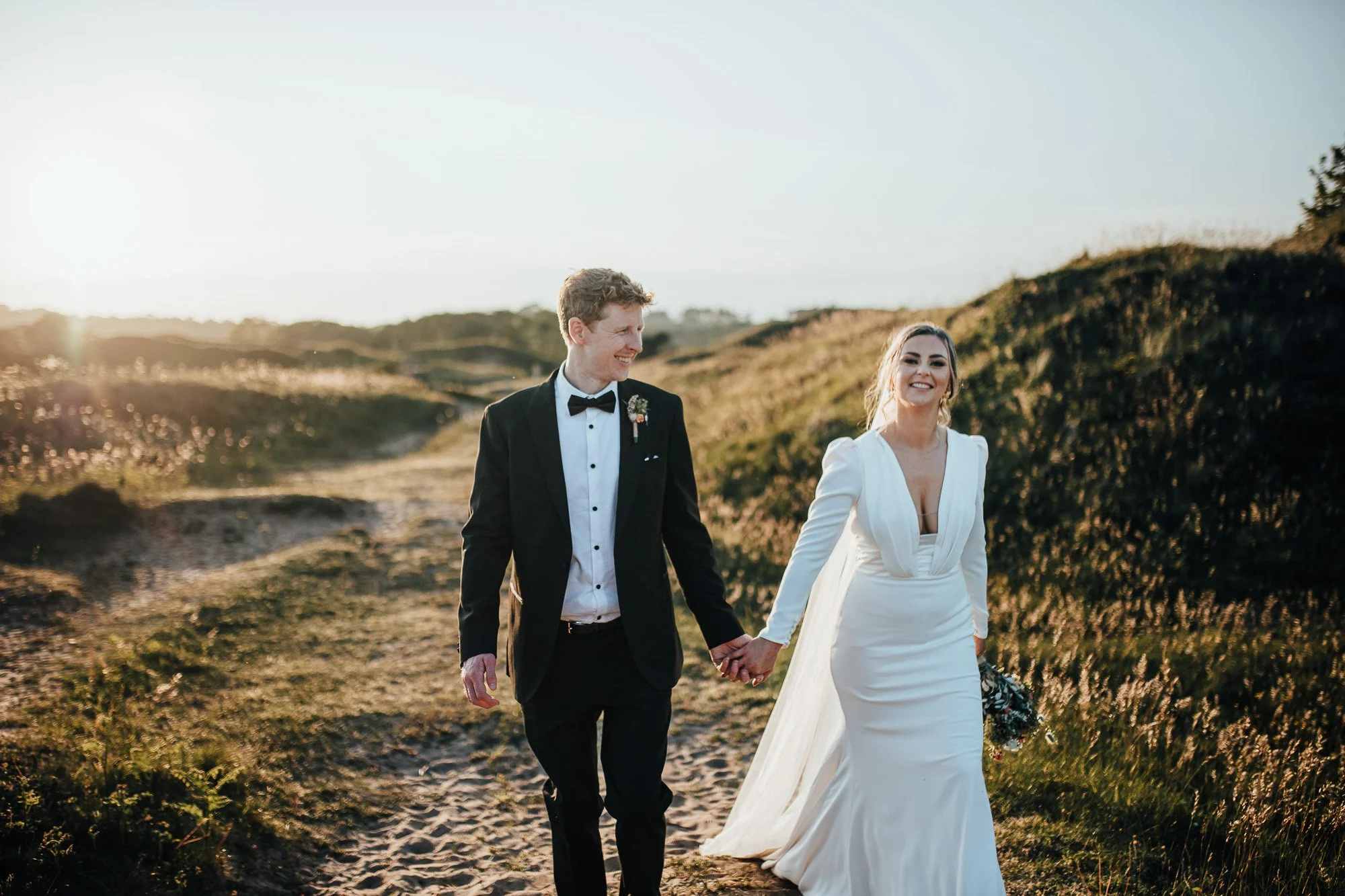 Bride and groom holding hands and walking on a grassy path during sunset, with the bride in a white dress and the groom in a black suit.