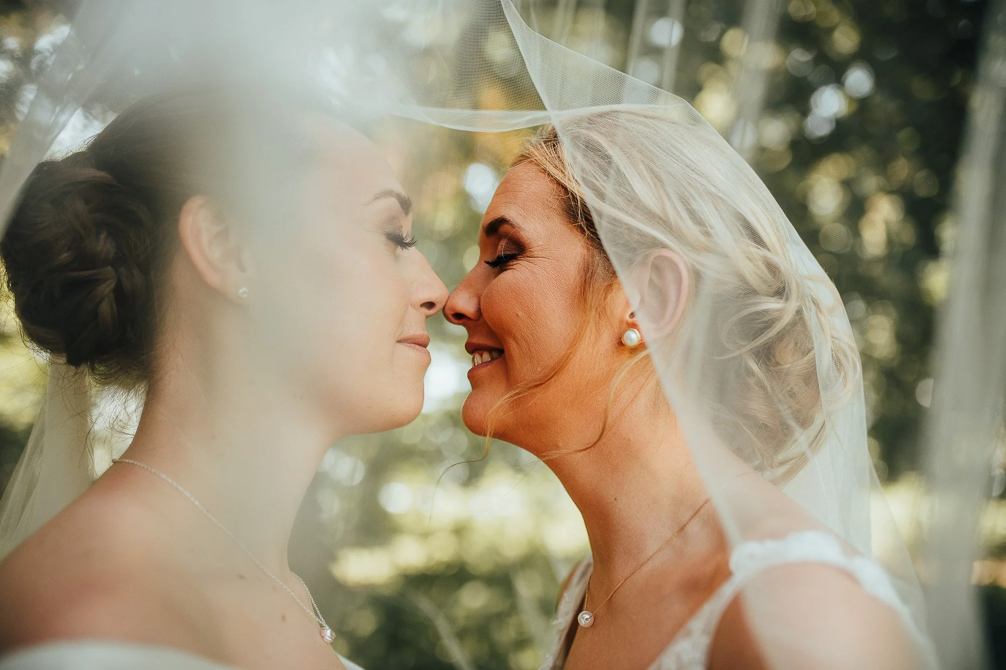 Two brides in wedding dresses under a veil, smiling face-to-face.