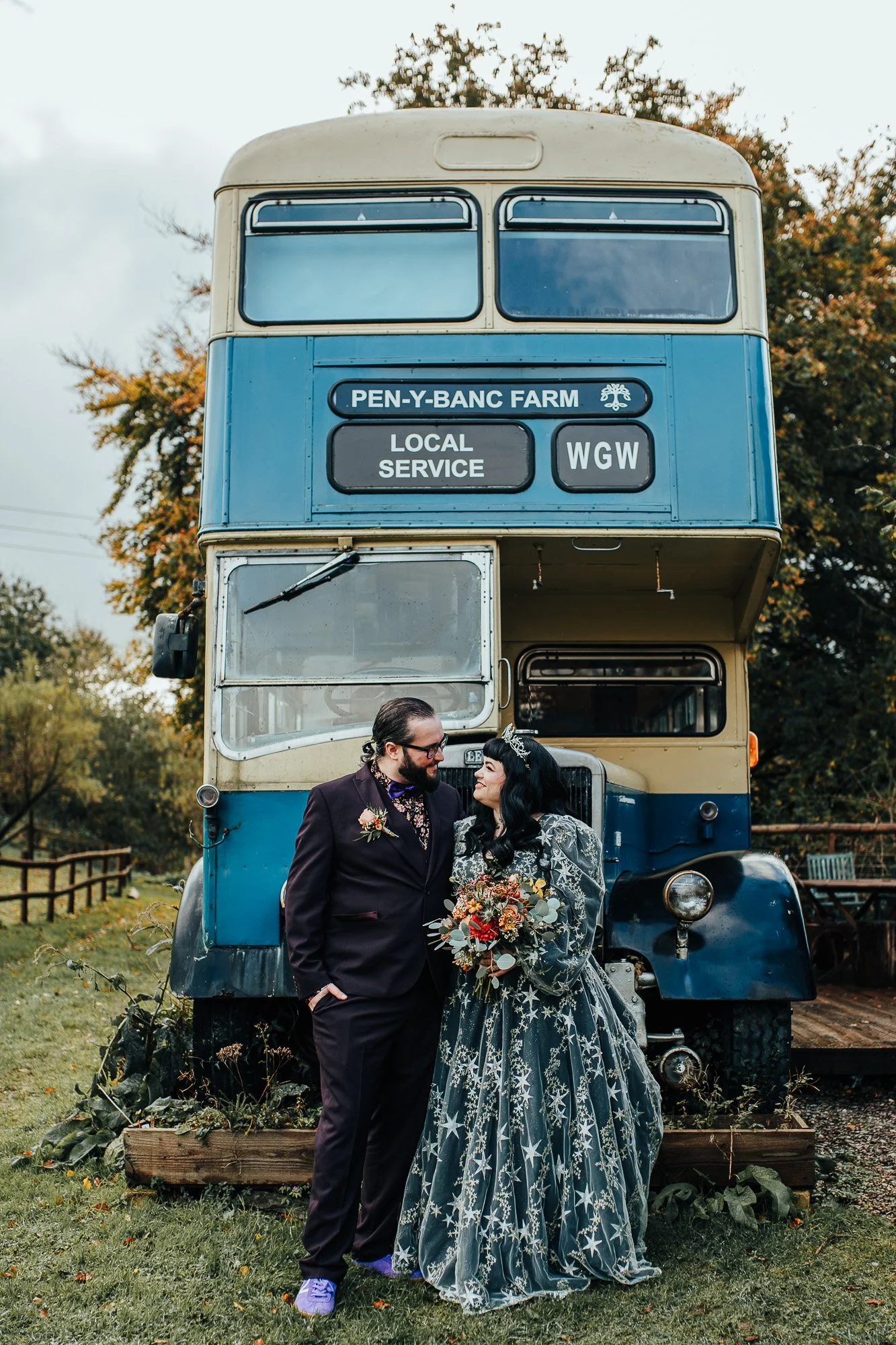 Couple in wedding attire standing in front of a vintage double-decker bus labeled 'Pen-y-Banc Farm Local Service WGW'.