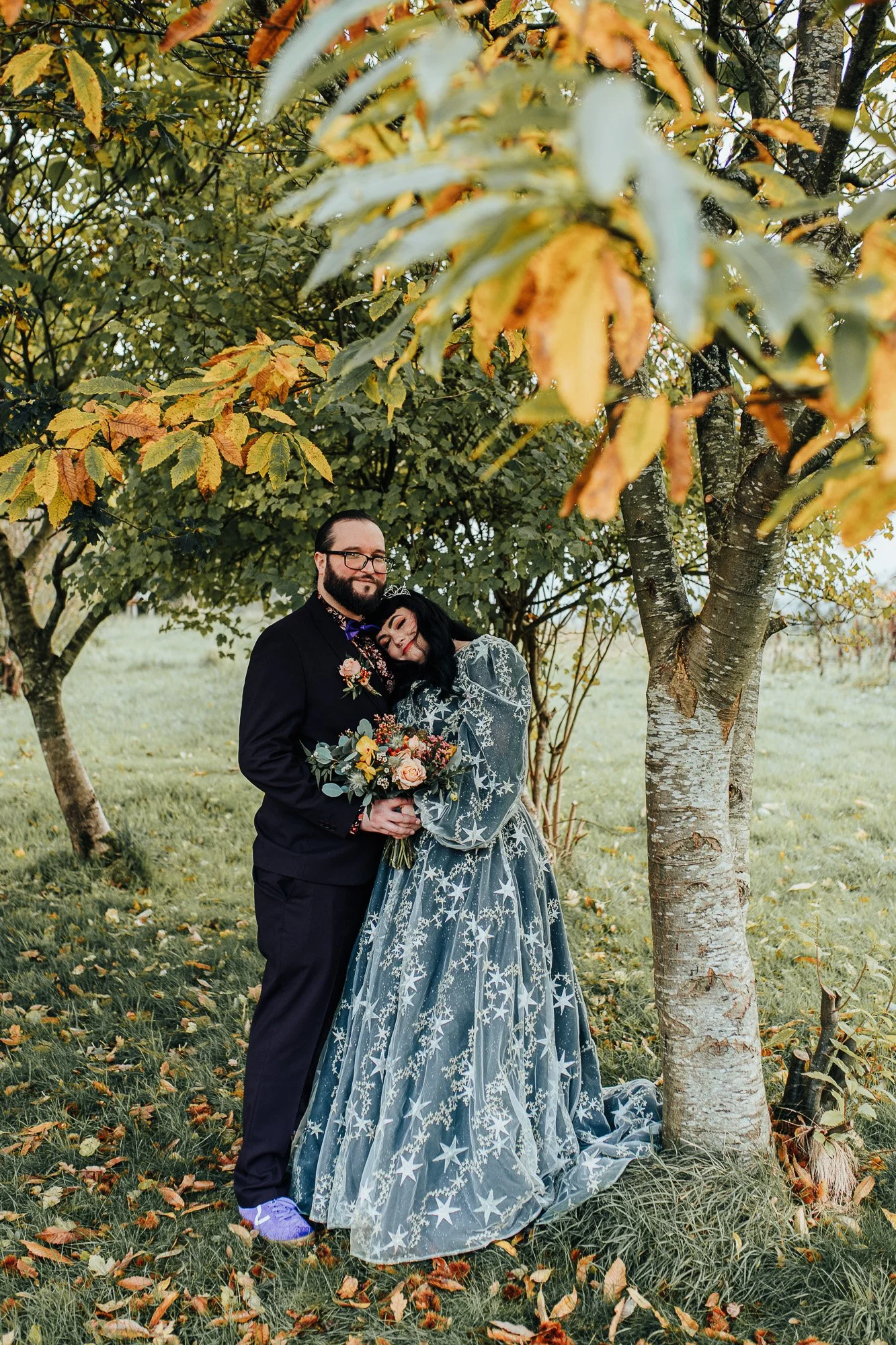 A couple embraces under a tree with autumn leaves. The woman is wearing a star-patterned dress and holding a bouquet.