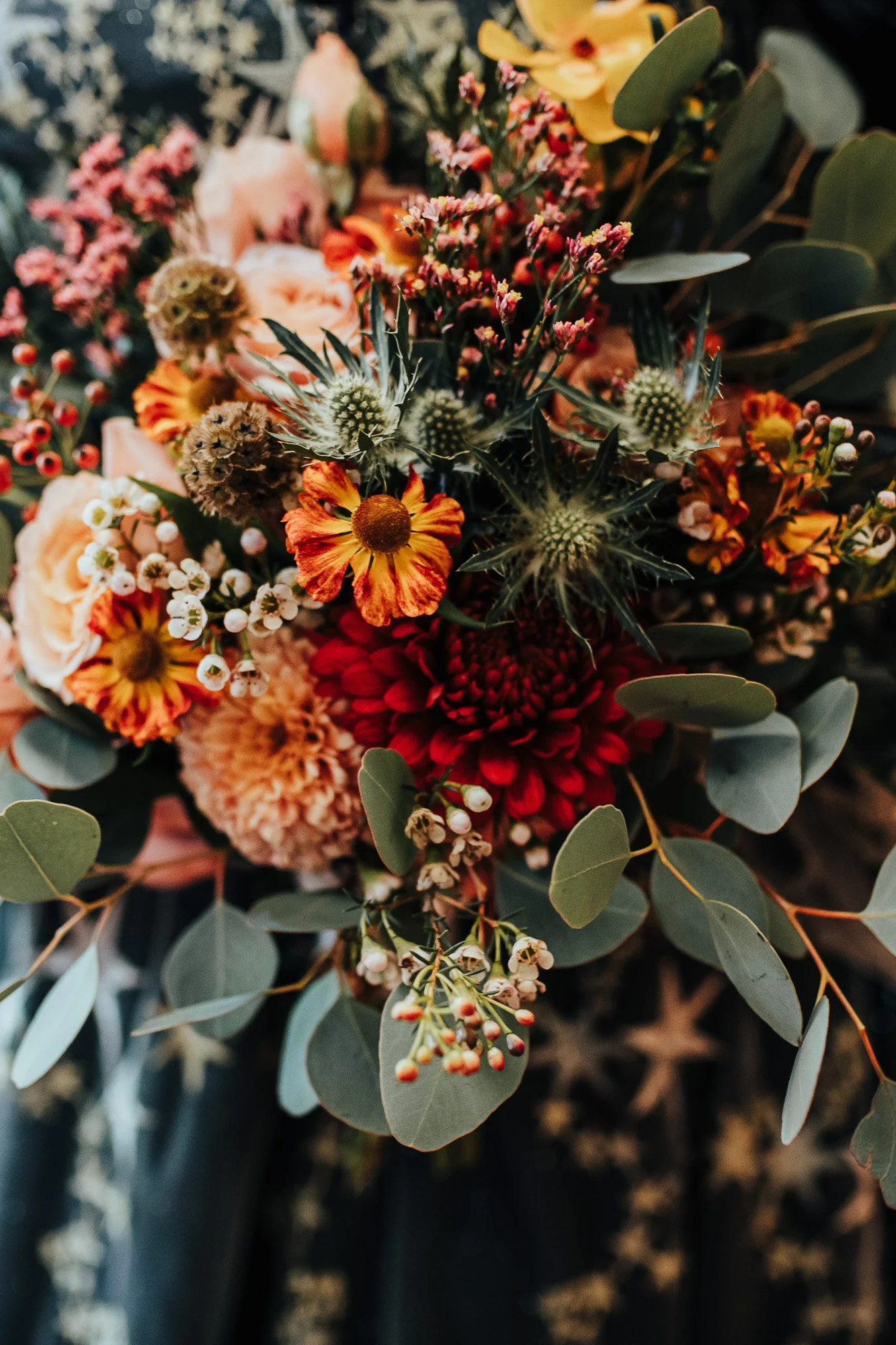 Colorful flower bouquet with orange, red, and yellow blooms, including roses and chrysanthemums, accented with green foliage and small white flowers.