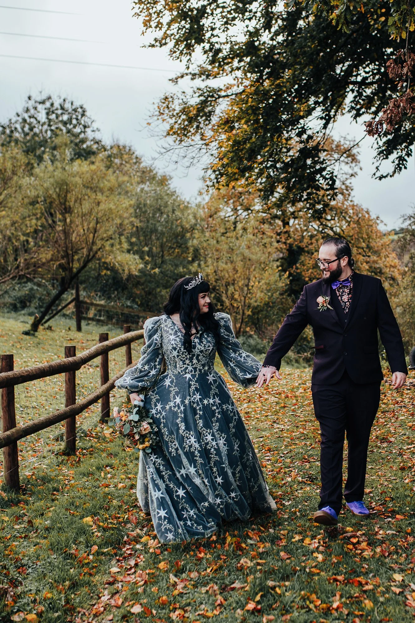 A couple in formal attire walking hand in hand on a grassy path. The woman is wearing a blue dress with star patterns and holding a bouquet, while the man is in a dark suit with a floral shirt. They are outdoors in a park-like setting with trees and 