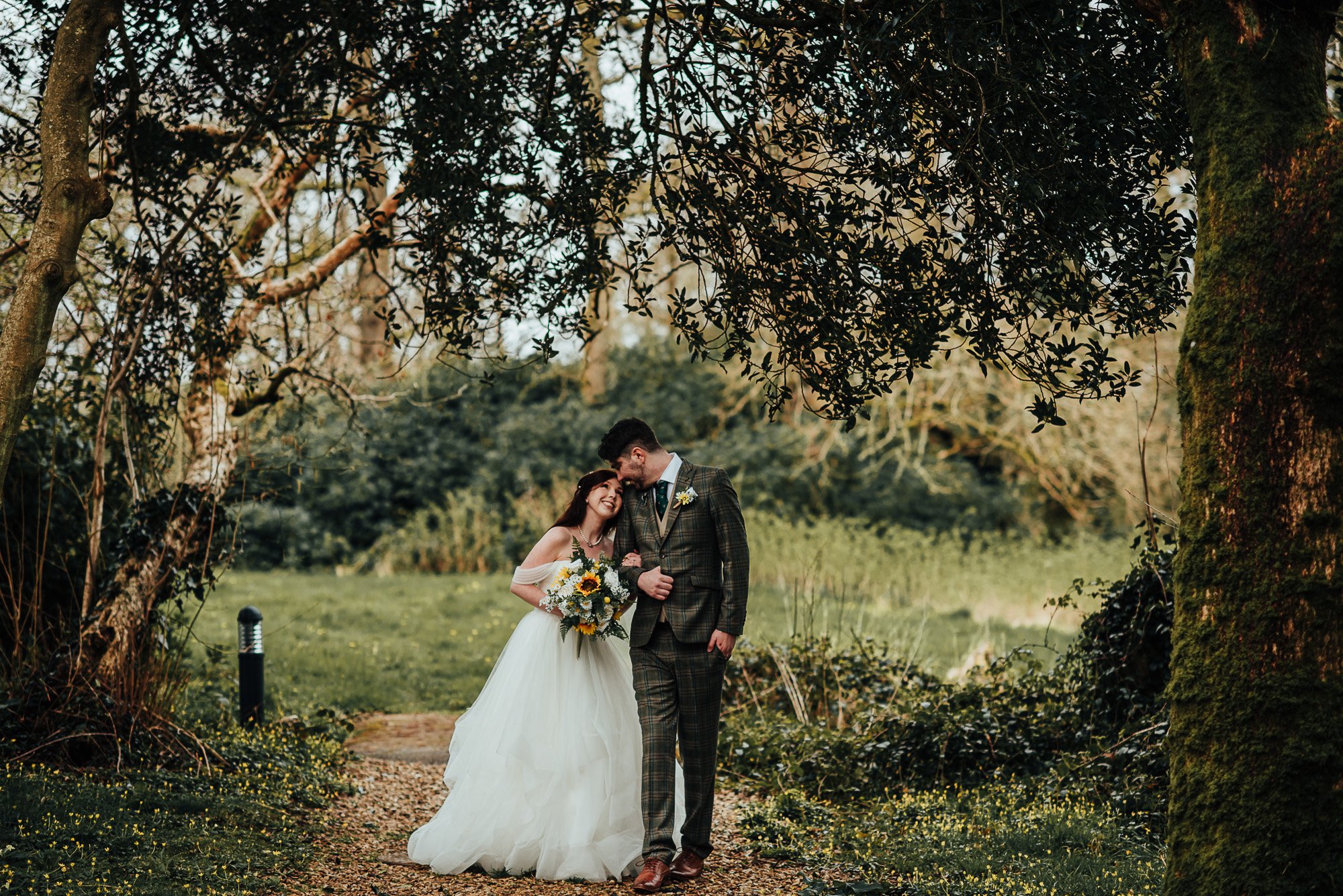 Bride and groom smiling and embracing in a forest setting, with the bride holding a bouquet and wearing a white dress, while the groom wears a plaid suit.